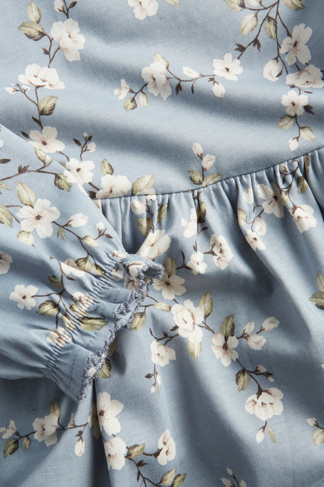 Zbliżenie jasnoniebieskiej kwiecistej sukienki dżersejowej z marszczonym materiałem i mankietem z gumką wykończonym picot.