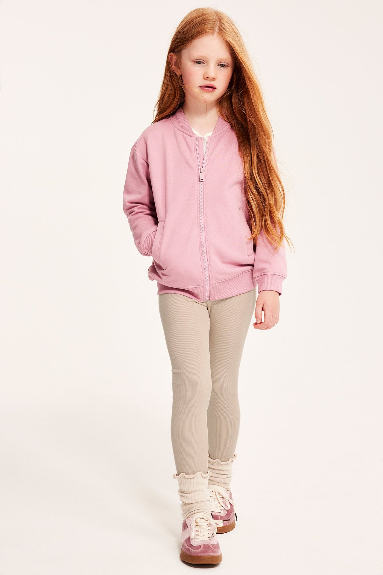 Edestä kuvattu lapsi beigessä ribbileggingsissä, vaaleanpunaisessa bomber-takissa, valkoisissa röyhelösukissa ja vaaleanpunaisissa lenkkareissa.