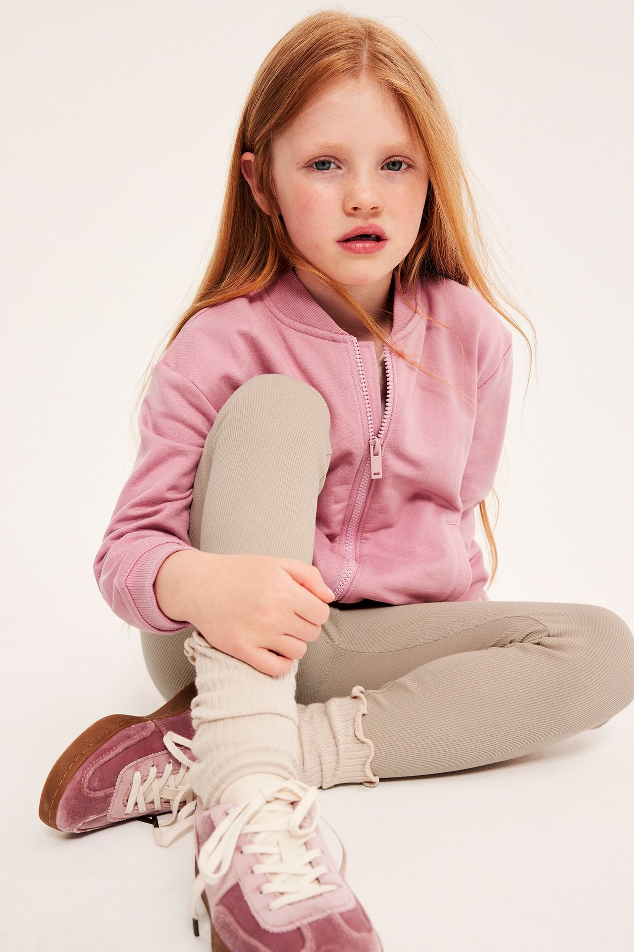 Tyttö vaaleanpunaisessa bomberjackassa ja beigeissä legginsissä, istuu lattialla.