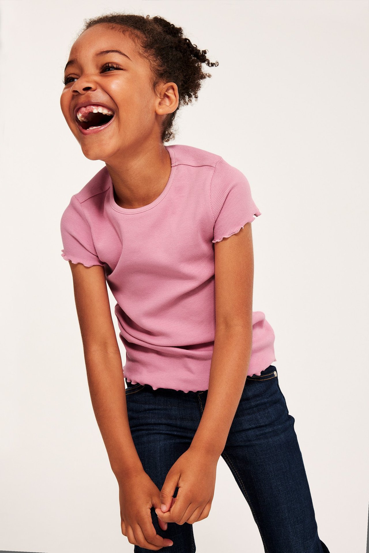 Framifrån, ett skrattande barn i en rosa ribbad kortärmad topp med babylock-kanter och mörkblå jeans.