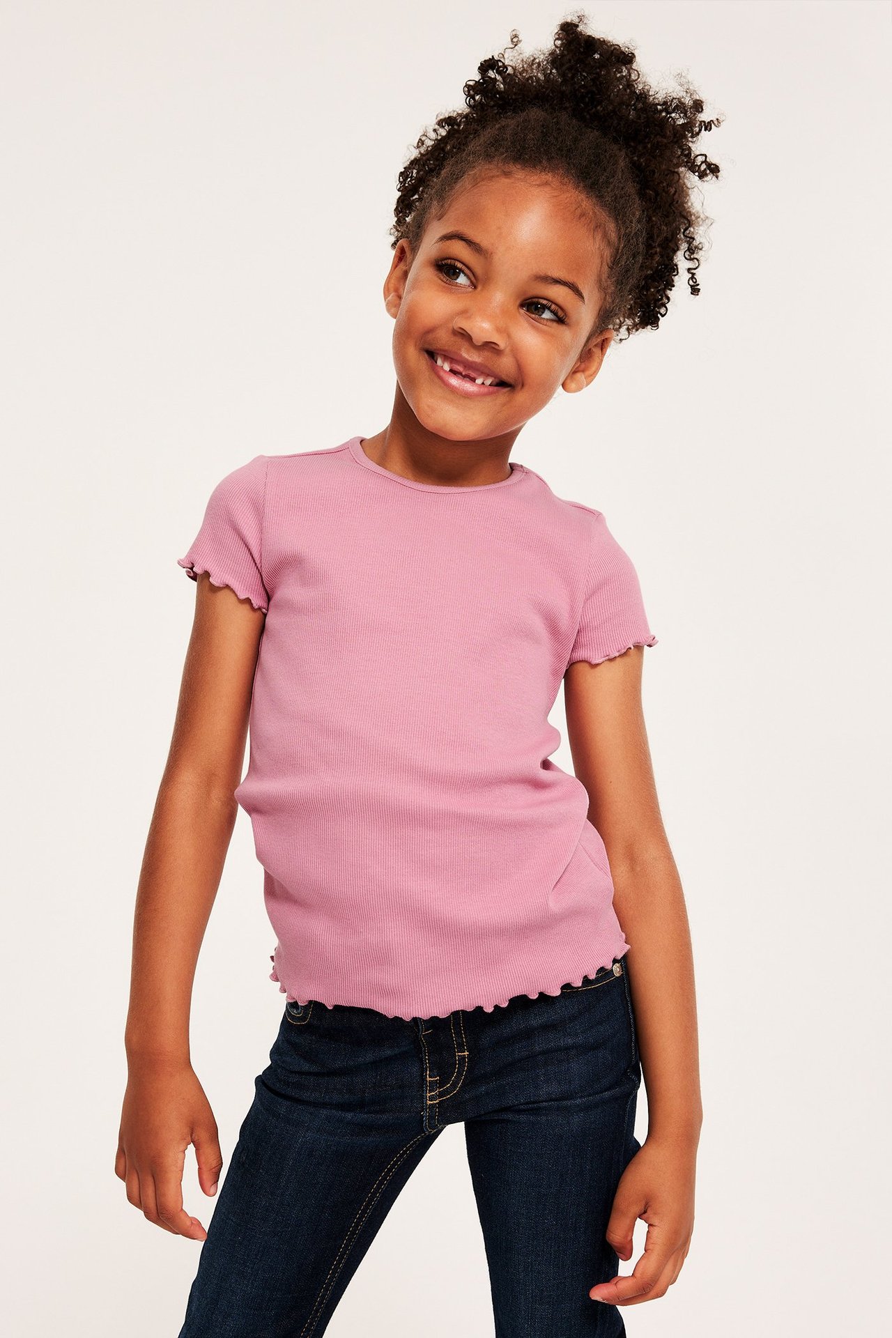 Frontvy av ett barn i en rosa ribbad kortärmad topp med babylock-kanter och mörkblå jeans.