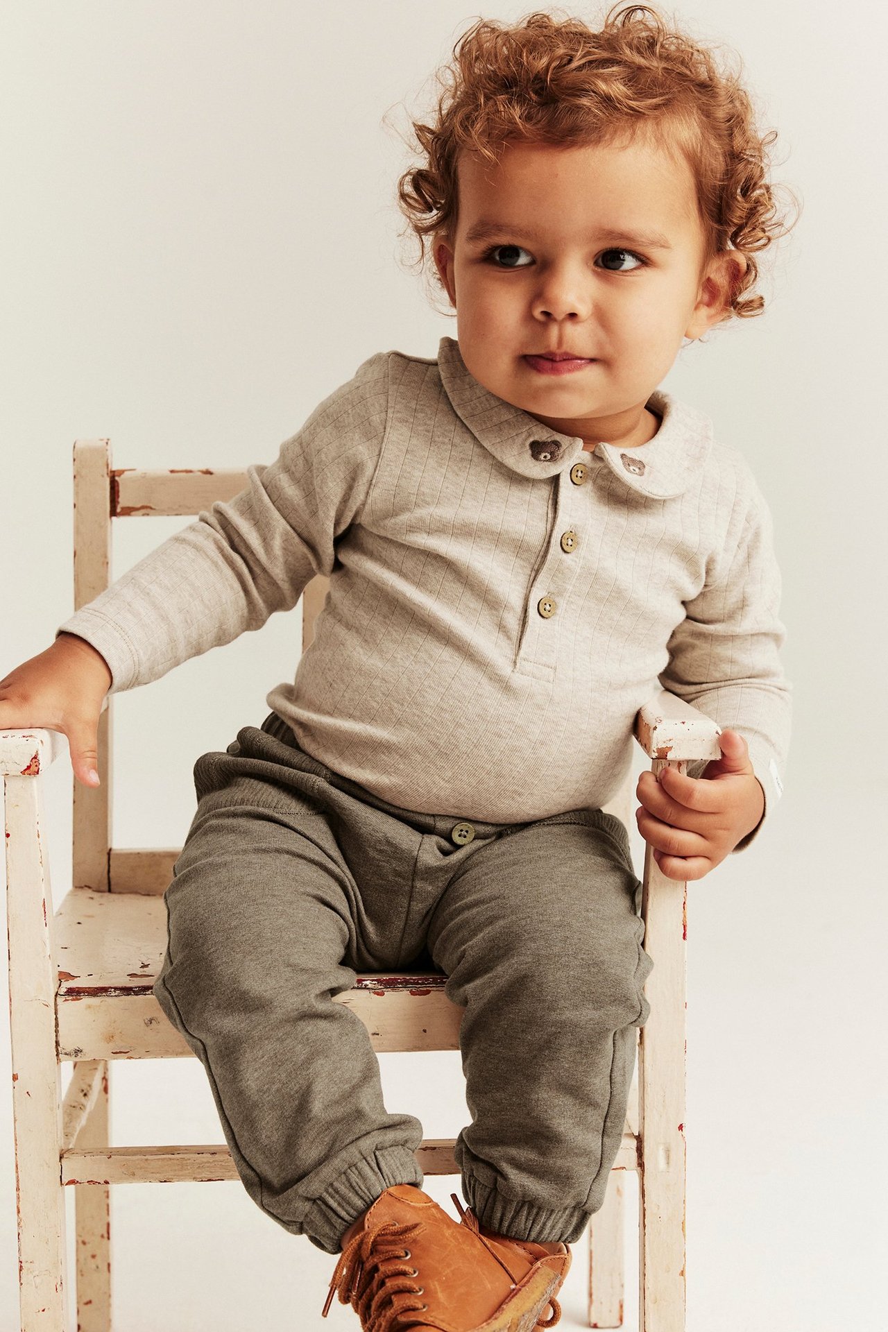 Widok z przodu: Niemowlę w brązowych spodniach dresowych z guzikami, jasnoszarym prążkowanym topie z kołnierzykiem i misiem oraz brązowych butach.