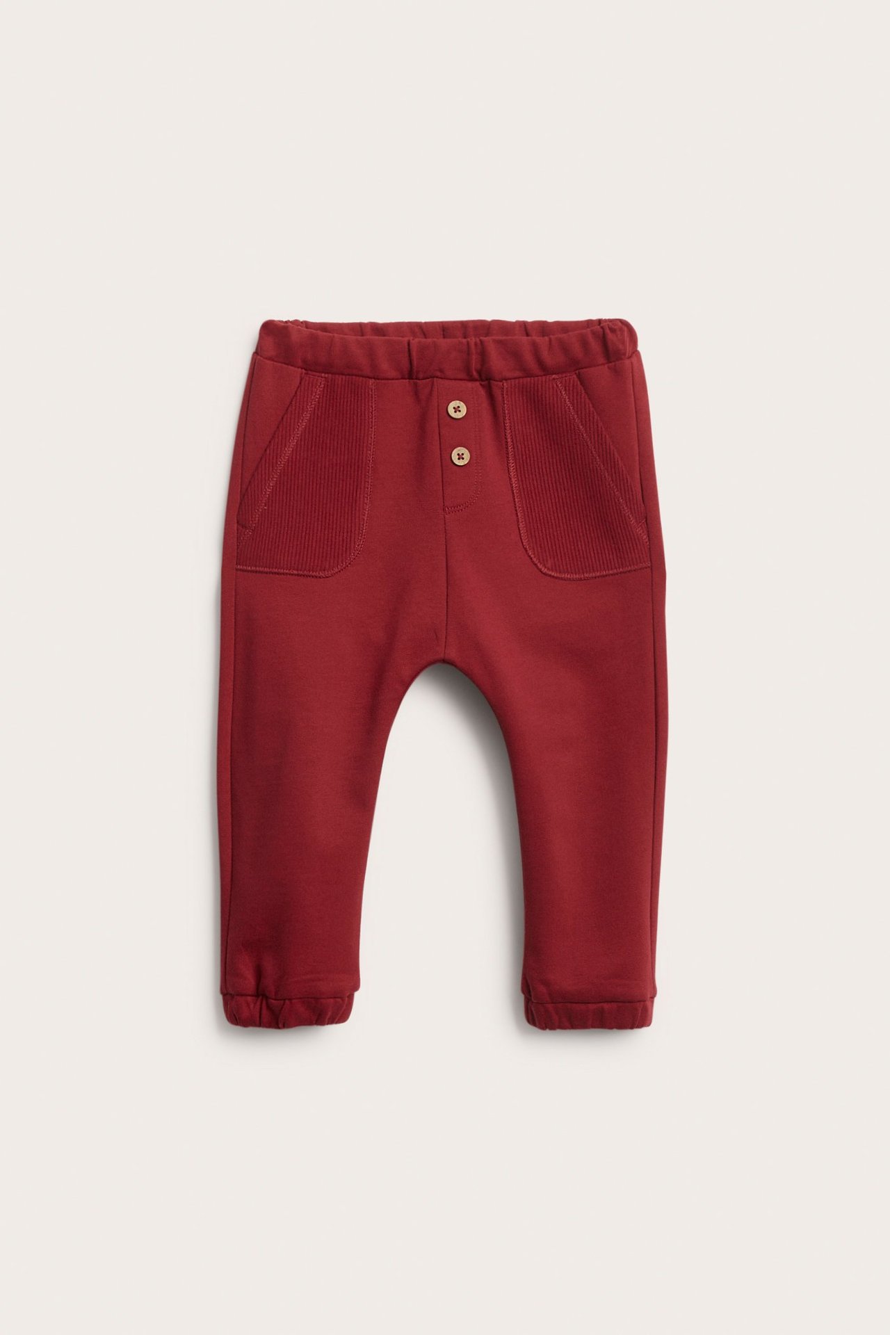 Frontvisning av røde babyjoggebukser med elastisk midje, ribbestrikkede lommer og to dekorative knapper.