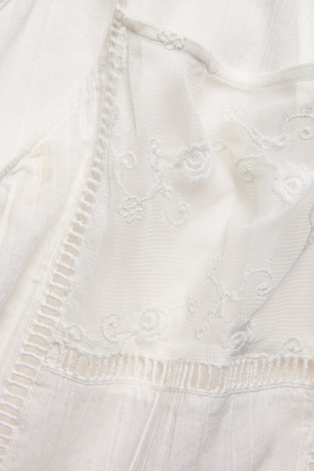 Zbliżenie na biały bawełniany top z delikatną koronką w kwiaty i haftowanymi detalami.