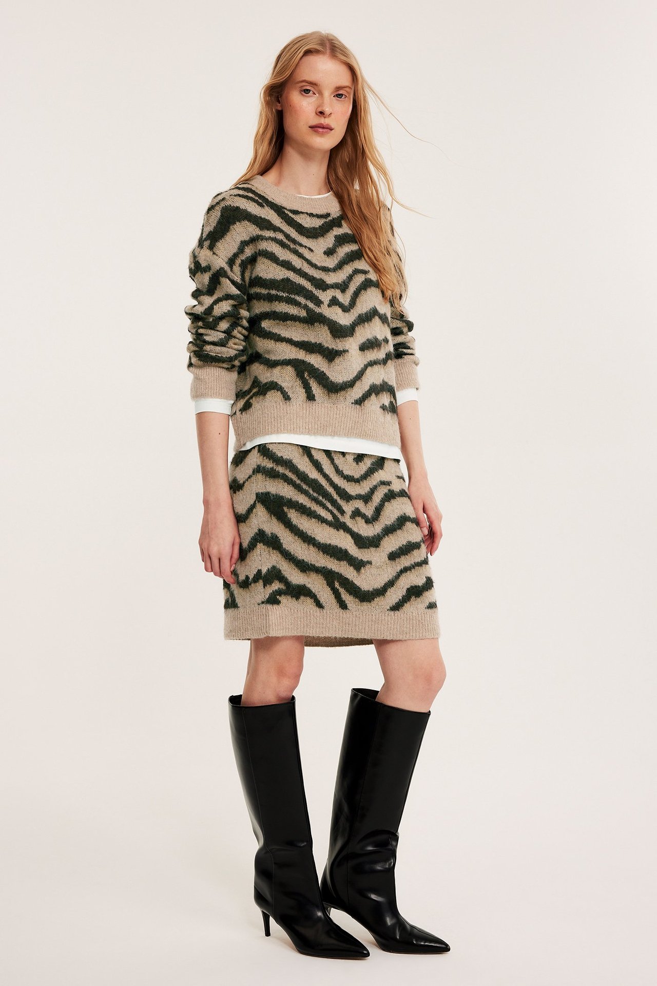 Widok z przodu: Kobieta w beżowej dzianinowej minispódniczce w zebrę, pasującym swetrze i czarnych kozakach.