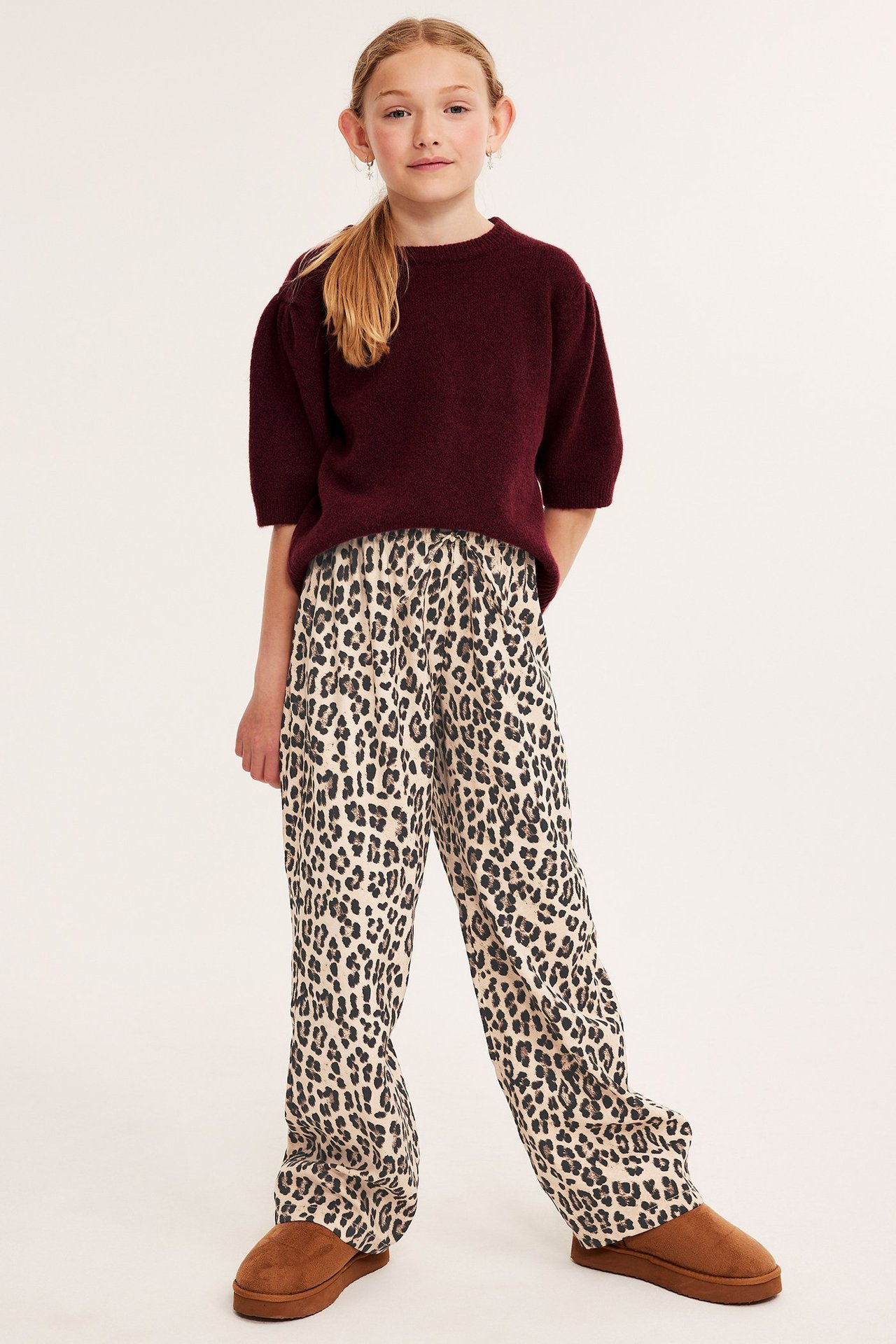 Edestä kuvattu lapsi yllään löysät beiget leopardikuvioiset housut, tummanpunainen neule ja ruskeat kengät.
