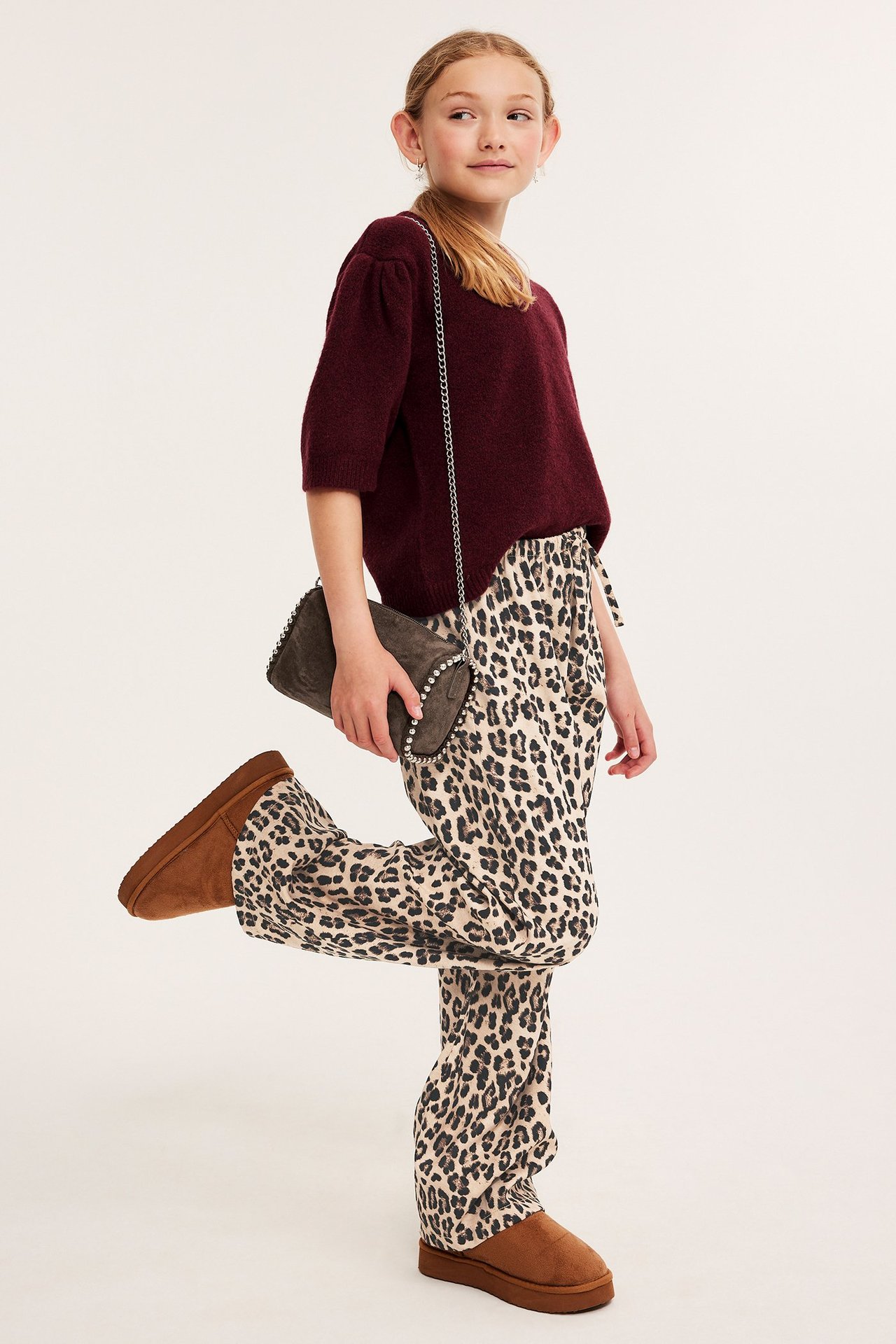 Widok z boku: Dziecko w luźnych brązowych spodniach w panterkę, bordowym swetrze, brązowych butach, trzymające szarą torebkę.