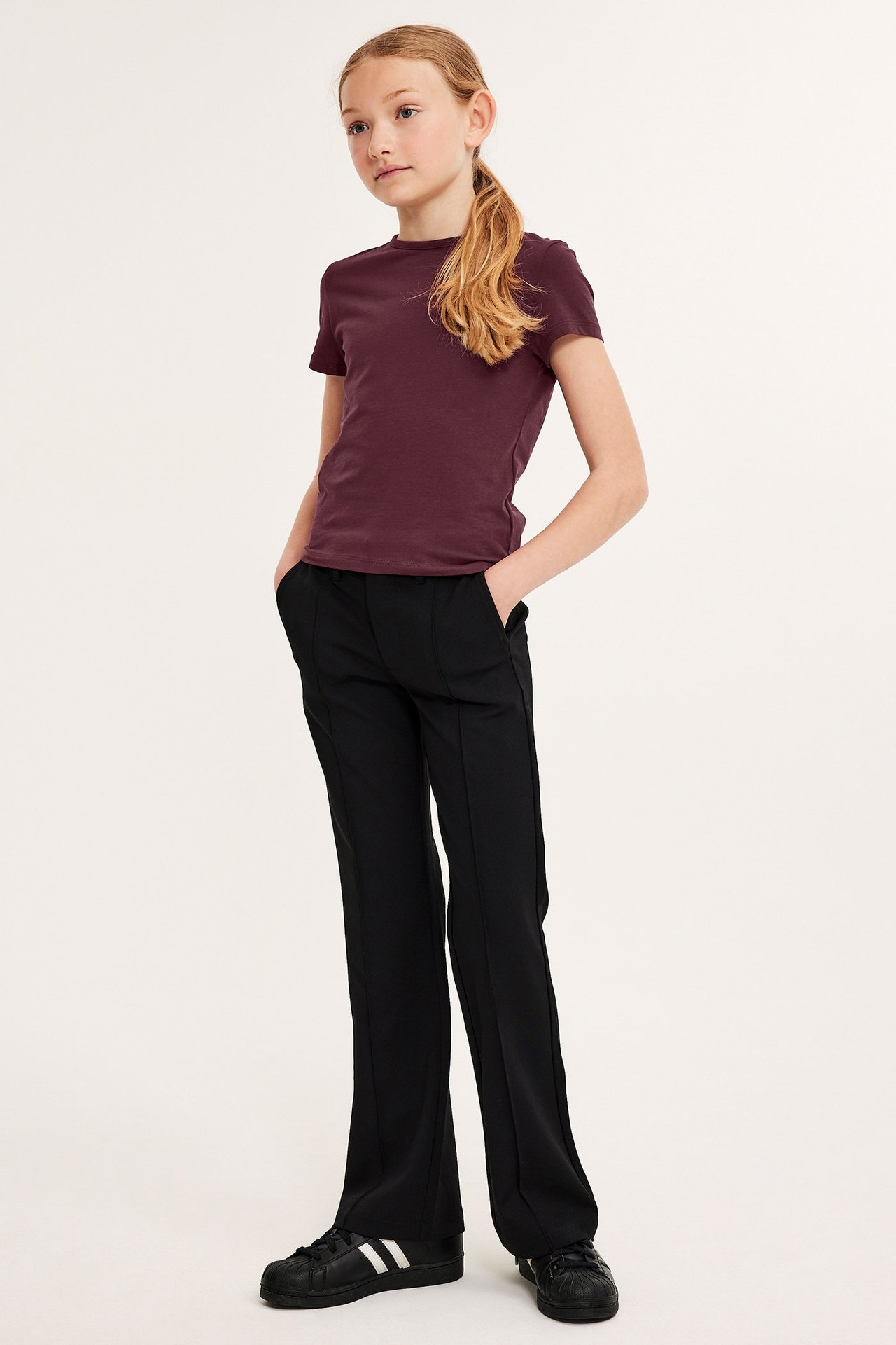 Helkroppsbild framifrån av ett barn i en mörkröd kortärmad åtsittande T-shirt, svarta byxor och svarta sneakers.