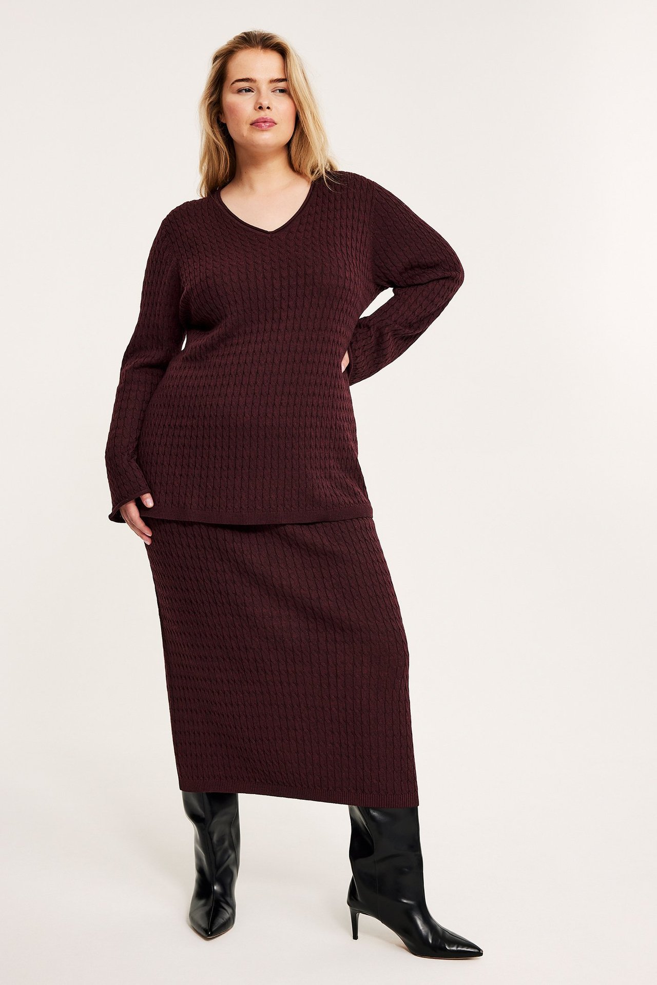 Forfra: Kvinne i vinrød kabelstrikket V-halsgenser, matchende skjørt og svarte støvler.