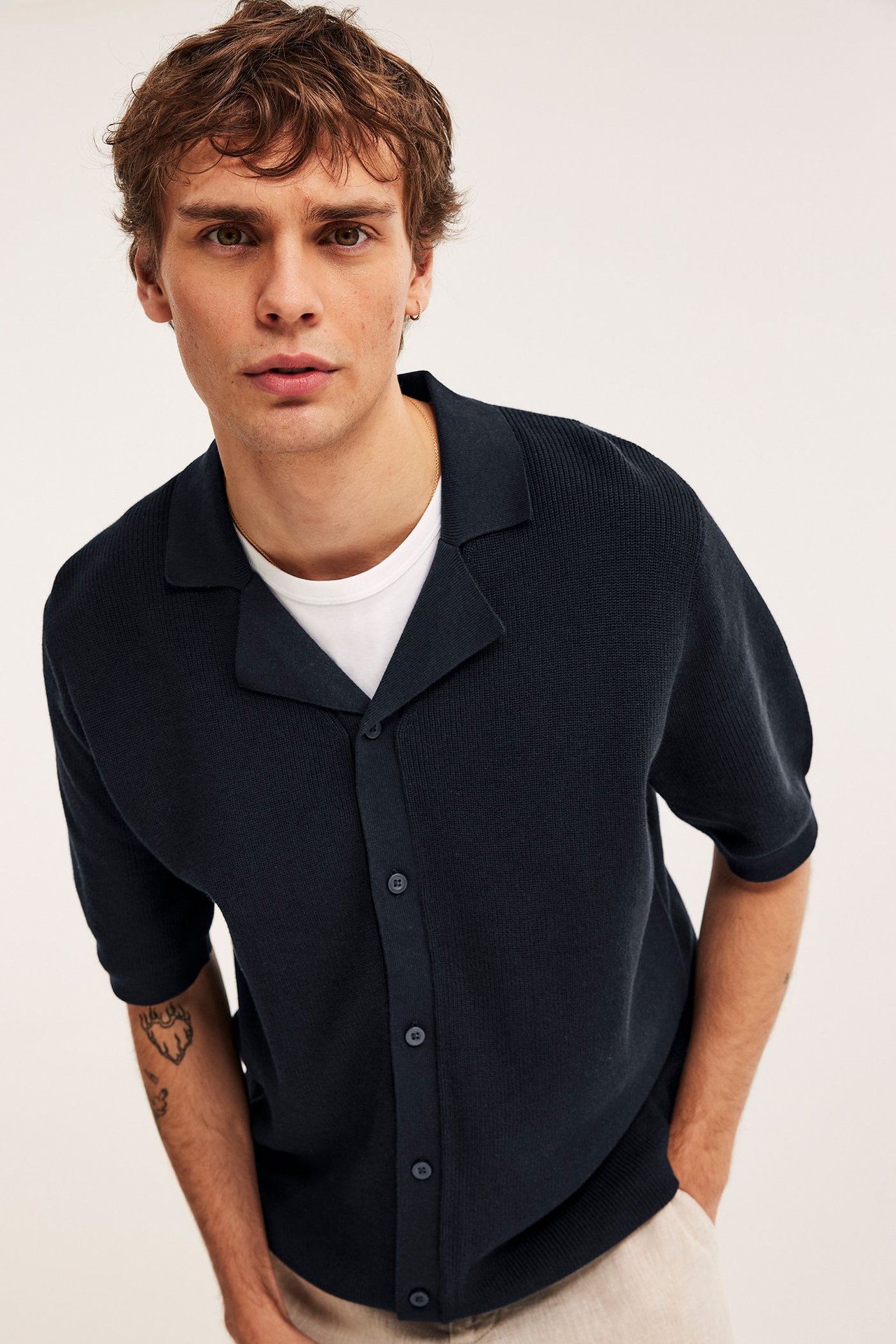 Mies edestä: tummansininen lyhythihainen neulottu pikeepaita valkoisen t-paidan ja vaaleanbeigen housujen päällä.