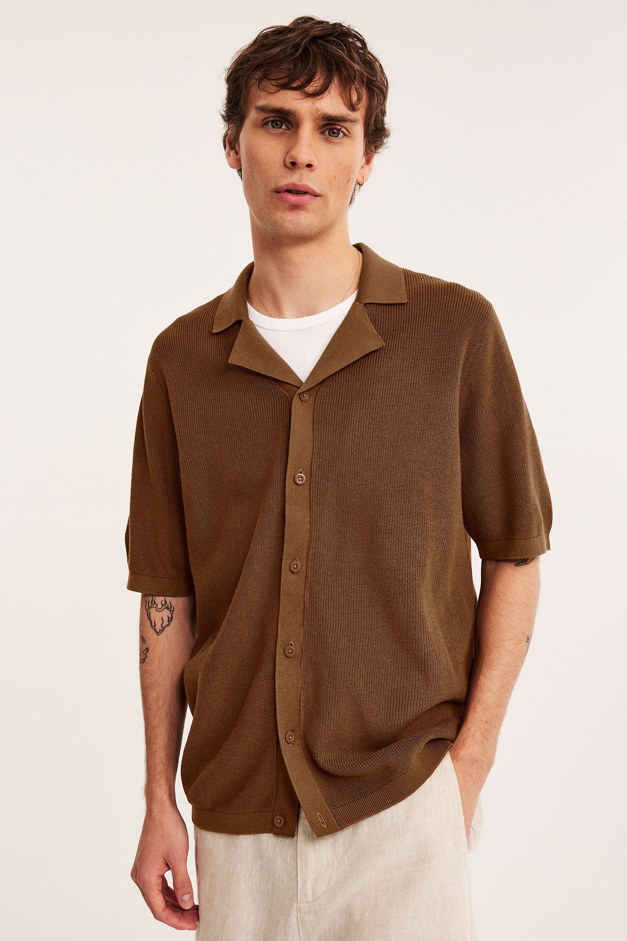 Edestä: mies ruskeassa puuvillaisessa neulotussa lyhythihaisessa pikeepaidassa, valkoisessa t-paidassa ja vaaleanbeigessä housuissa.