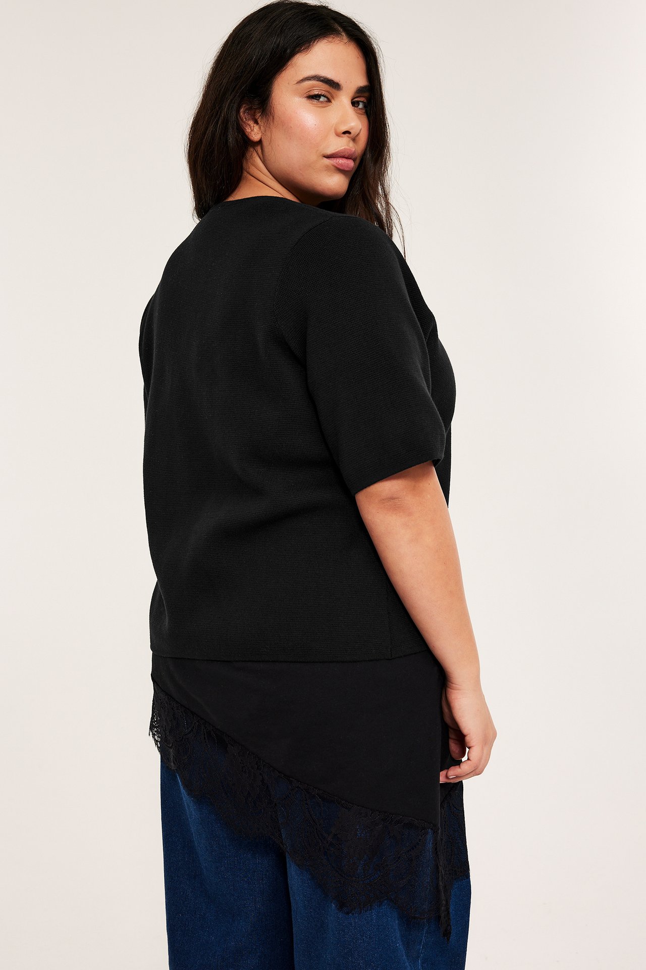 Naisen sivukuva yllään musta lyhythihainen neuletoppi epäsymmetrisellä pitsireunalla, sinisten farkkujen päällä.