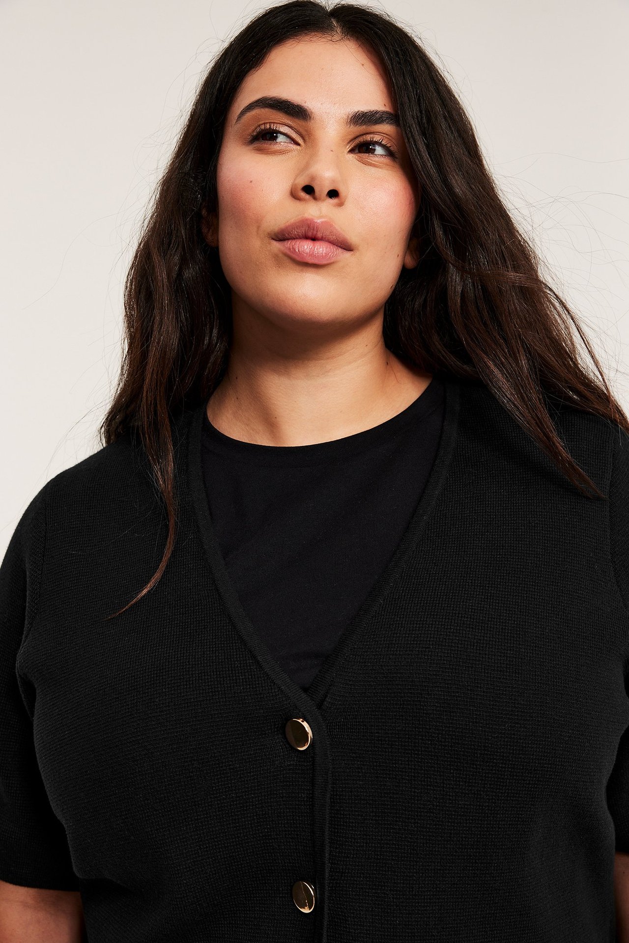 Närbild: kvinna i svart kortärmad V-ringad kofta med guldfärgade knappar över en svart topp.