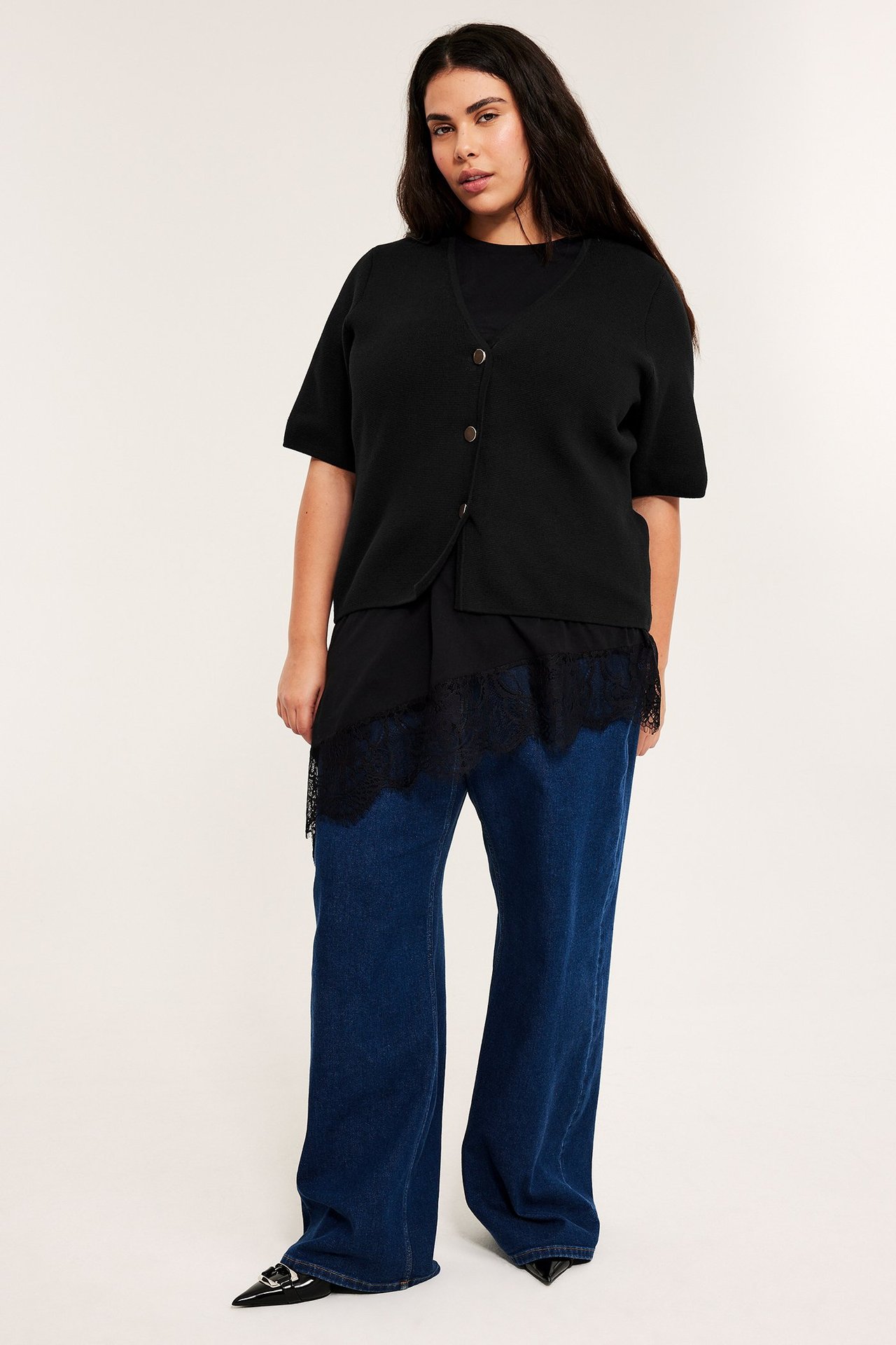 Framifrån: Kvinna bär svart kortärmad knapptopp, svart spetskantad topp, vida blå jeans, svarta skor.