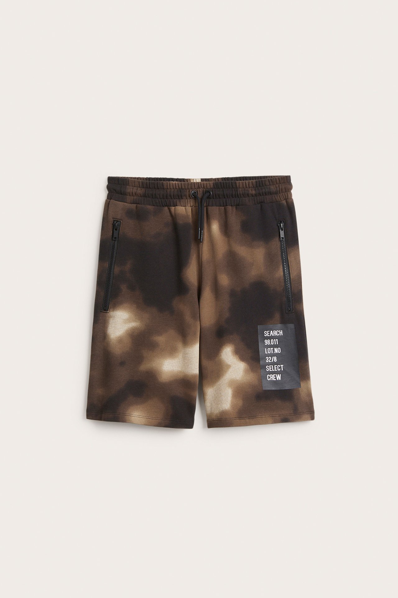 Widok z przodu dziecięcych brązowo-beżowych szortów tie-dye, ze sznurkiem, kieszeniami na zamek i naszywką.