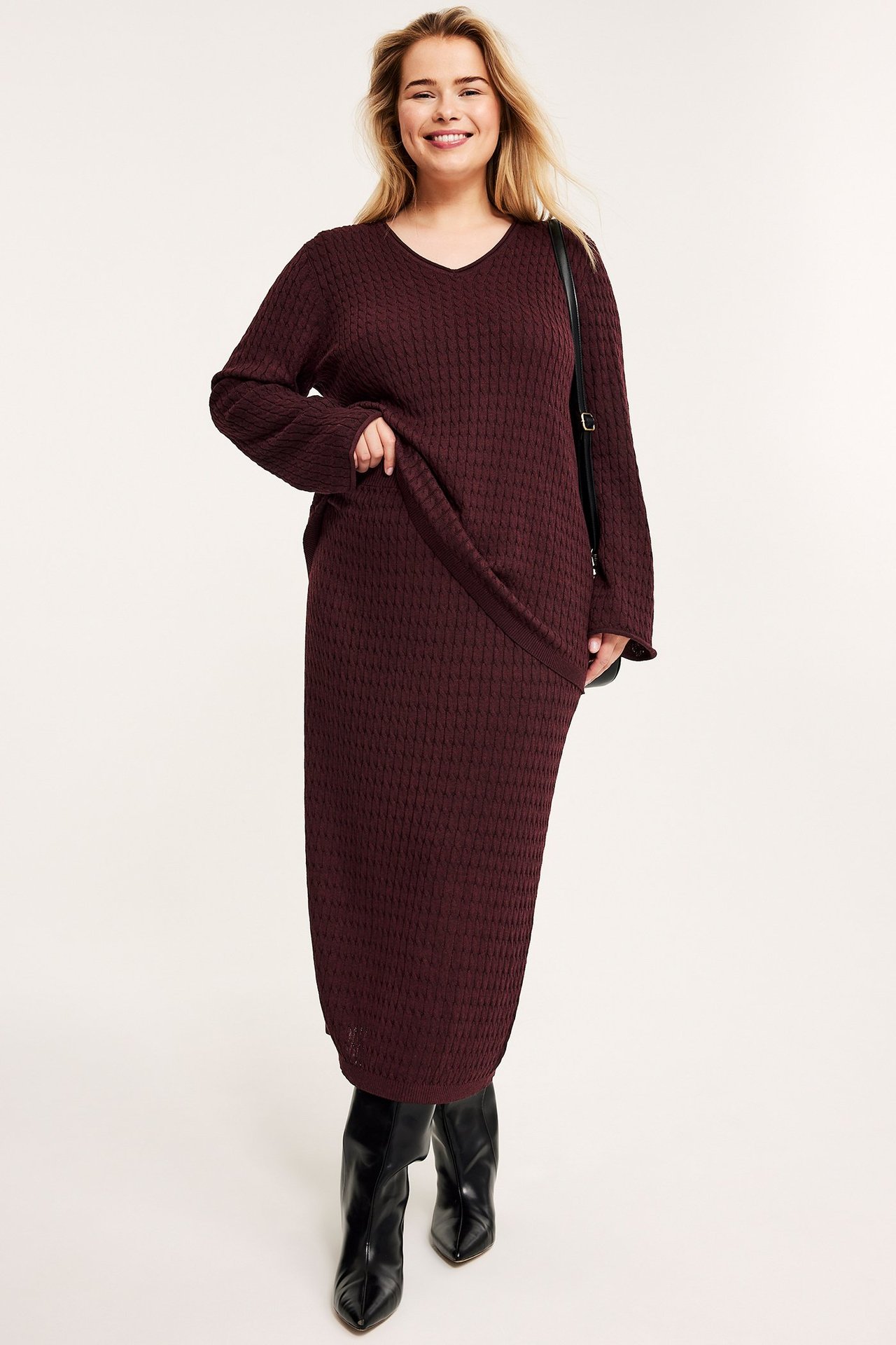 Forfra: kvinne i vinrød kabelstrikket midiskjørt og matchende genser, svarte støvler og skulderveske.