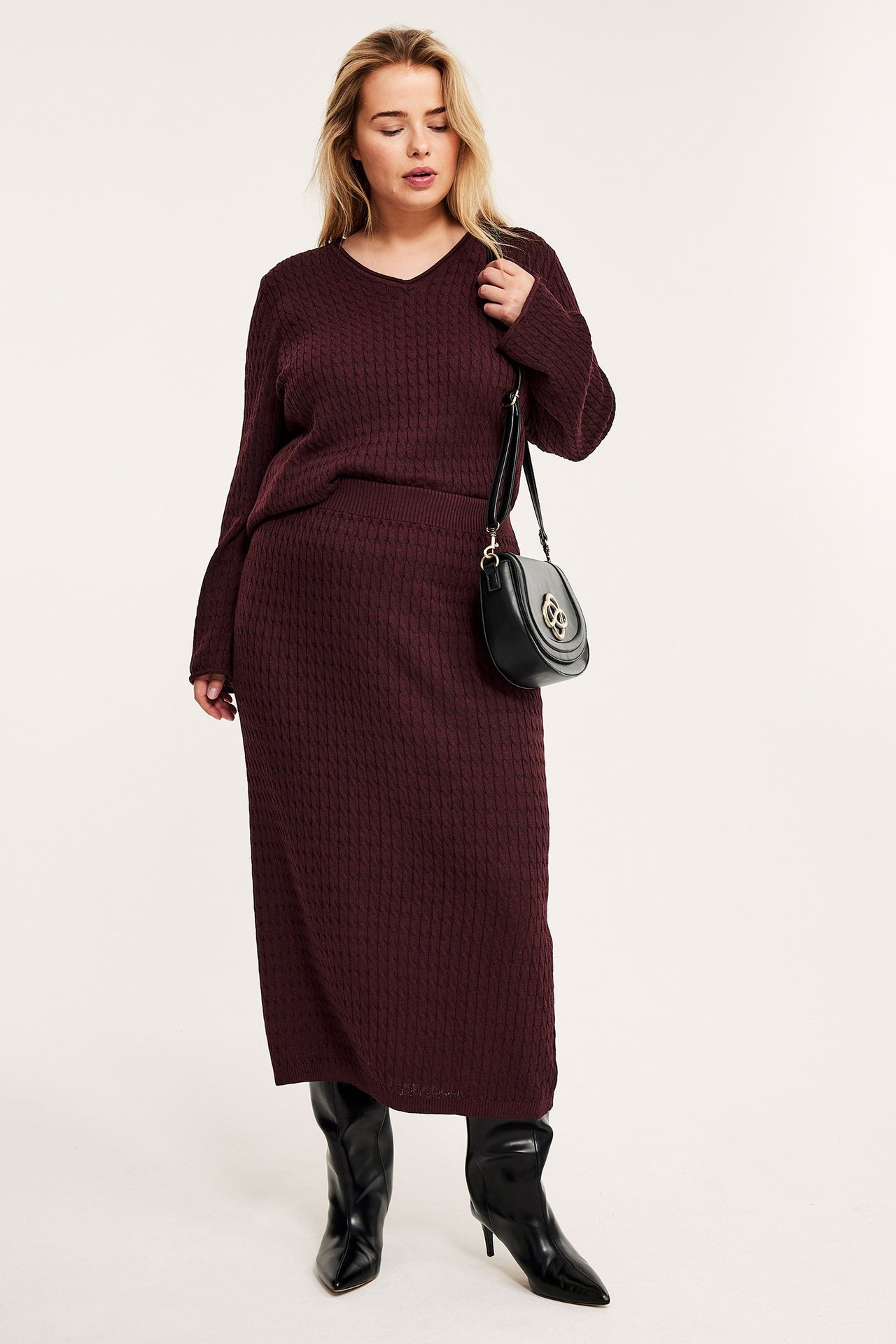 Widok z przodu: Kobieta w bordowej spódnicy midi z warkoczowym splotem, swetrze, czarnych butach, z czarną torebką.