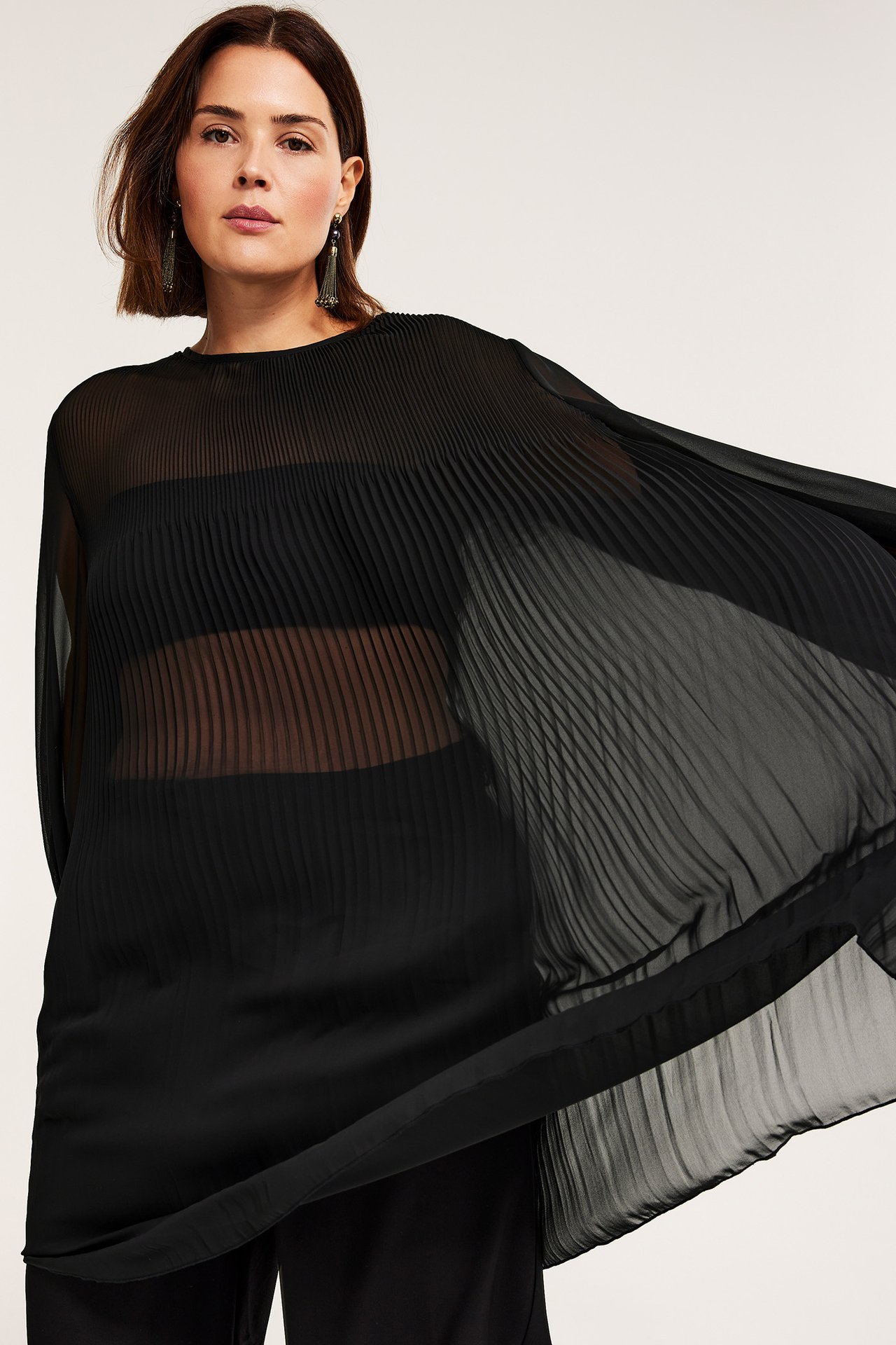 Framifrån, kvinna i skir svart plisserad polyesterklänning, stylad med svart BH och mörka örhängen.