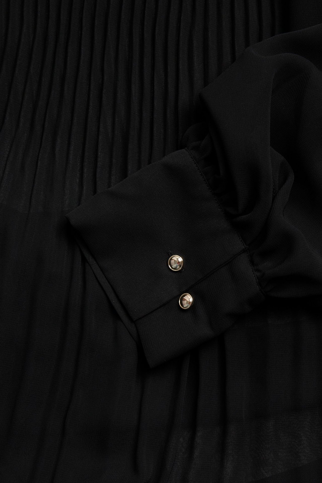 Zbliżenie czarnej plisowanej sukienki szyfonowej, mankiet rękawa z dwoma srebrnymi guzikami.