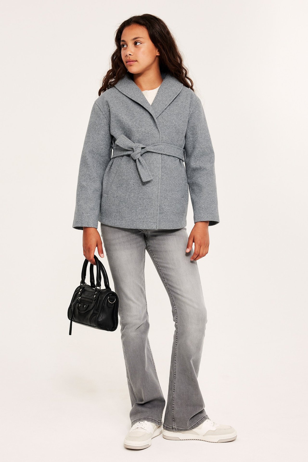 Frontvy: Flickbarn i ljusgrå melerad knytjacka, grå utsvängda jeans, vita sneakers, håller svart väska.