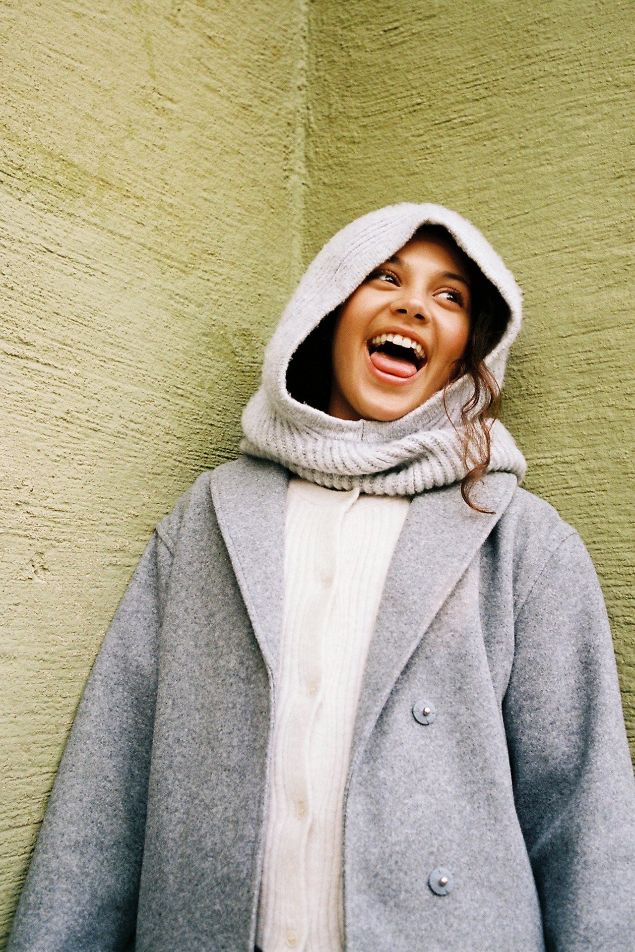 Widok z przodu: Kobieta w jasnoszarym melanżowym płaszczu, białym kardiganie i jasnoszarym dzianinowym kapturze.