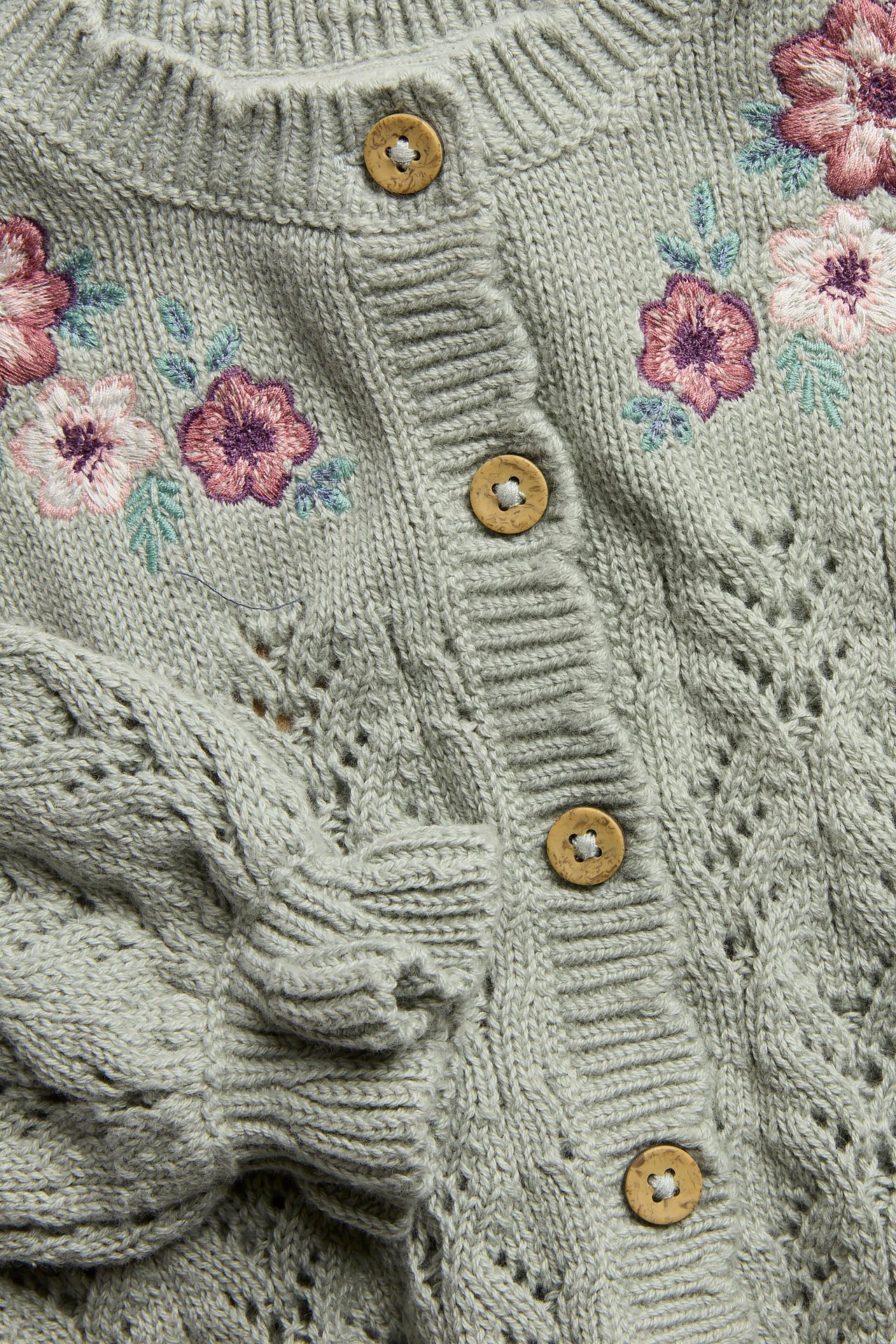 Närbild framifrån på en grön mönsterstickad bomullskofta med blombroderier och träknappar.