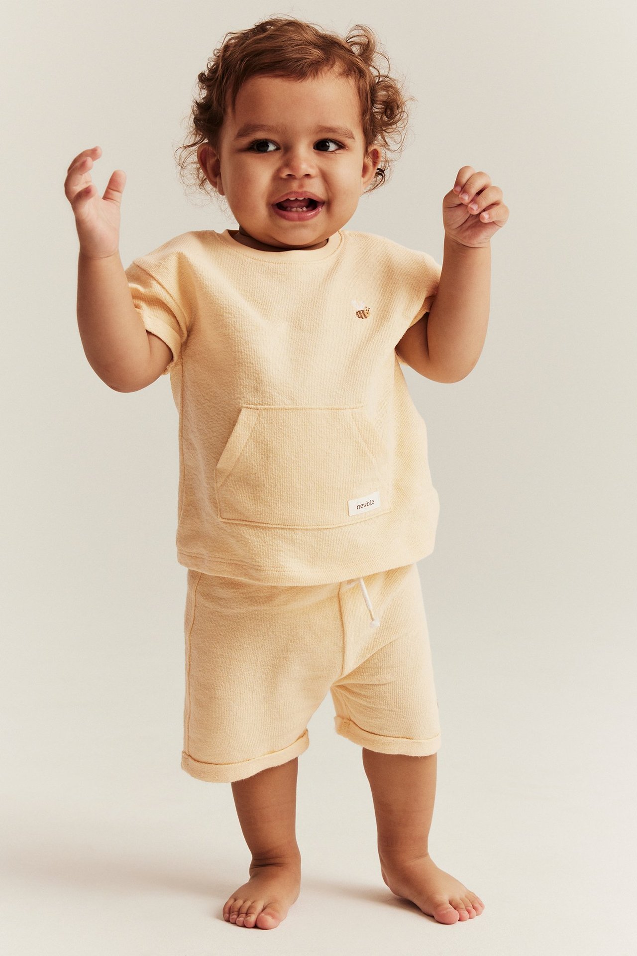 Edestä: vauva keltaisissa puuvillashortseissa, joissa kiristysnyöri, ja yhteensopivassa t-paidassa mehiläisbrodeeraus.