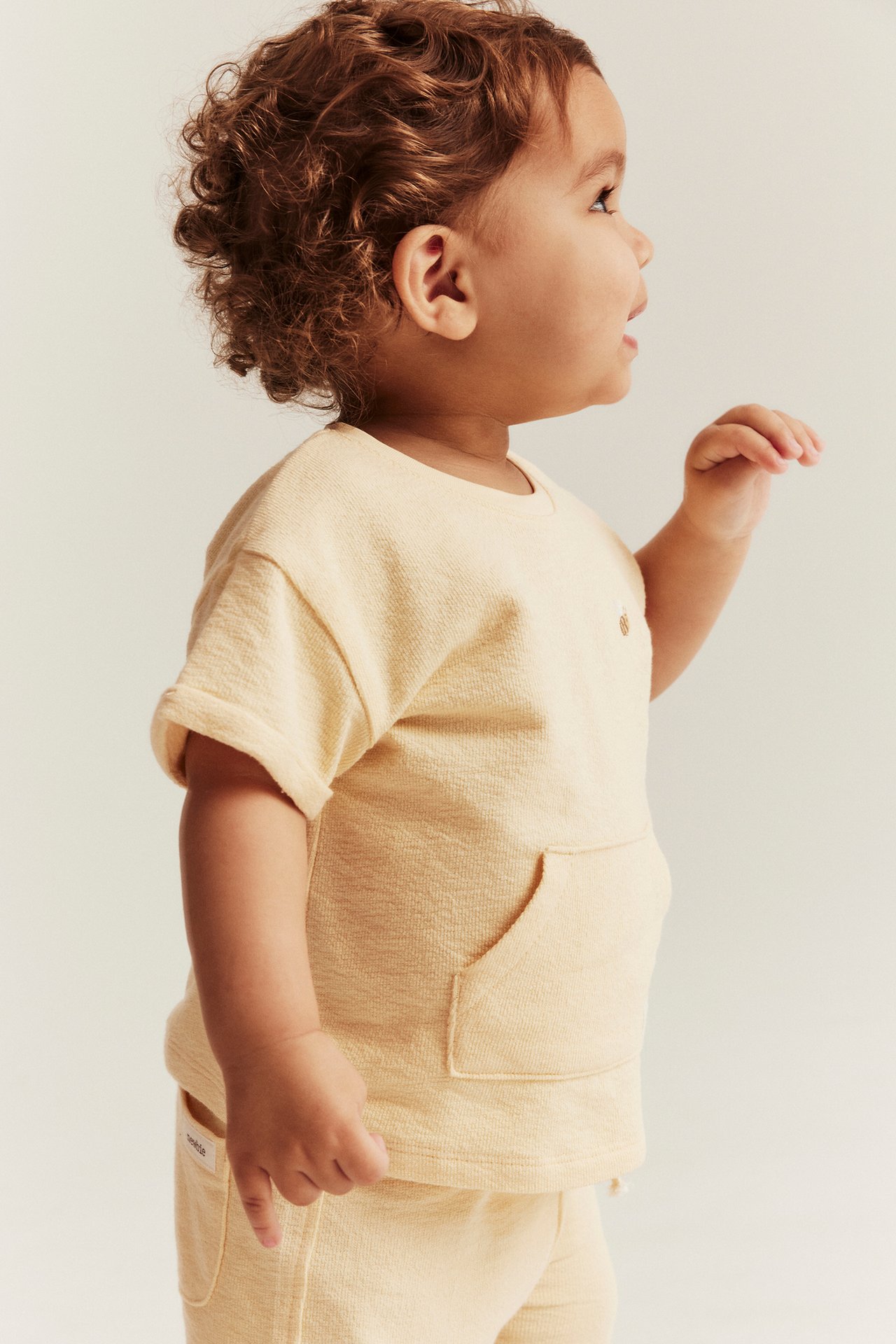 Sidebilde av en baby i en lys gul bomulls-t-skjorte med brystlomme og matchende shorts.