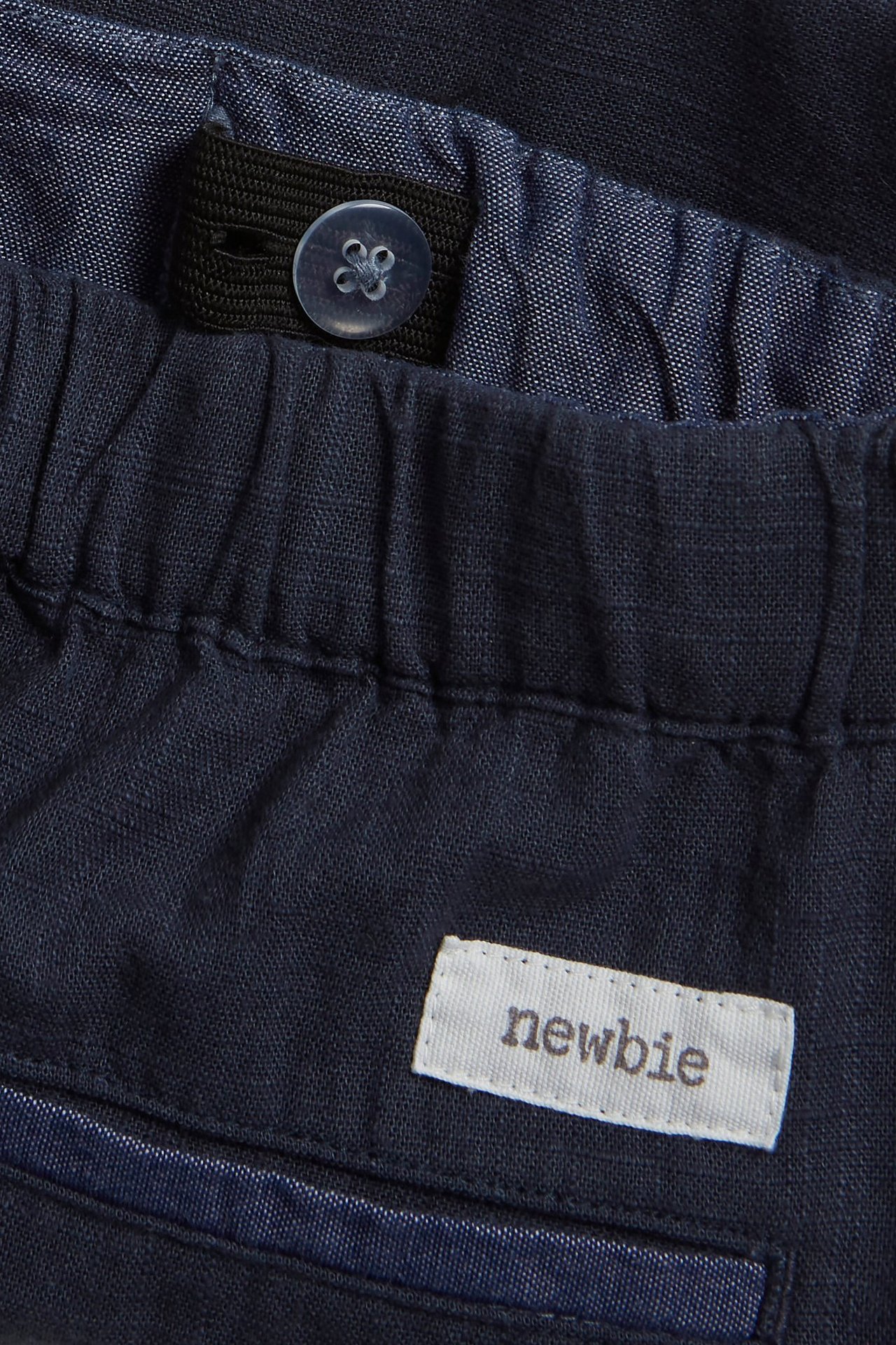 Närbild av mörkblå vävda shorts med resårlinning, justerbar innerresår, knapp och 'newbie'-etikett.