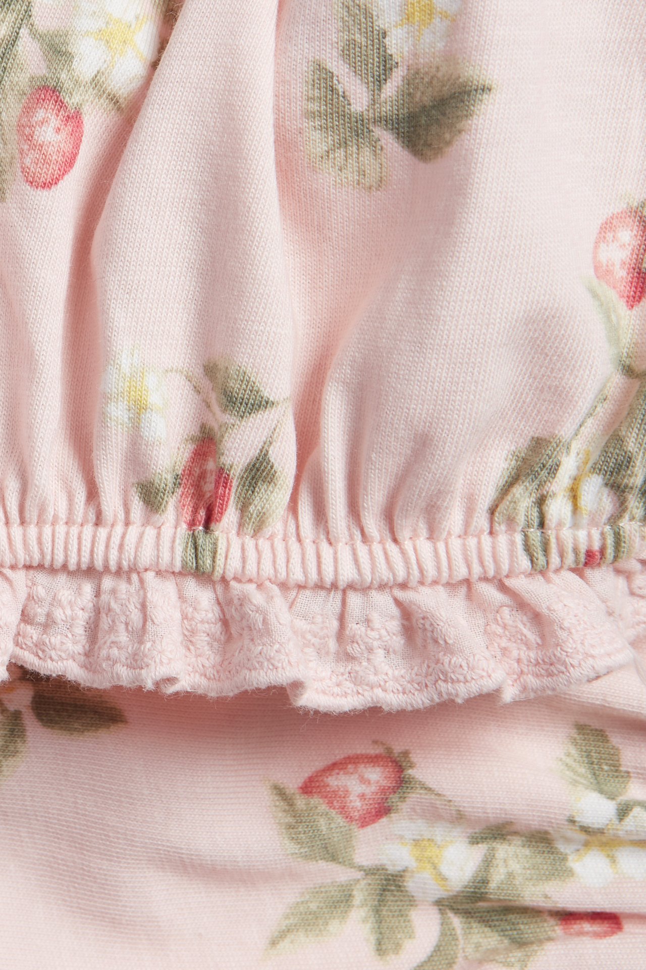 Nærbilde av lys rosa jersey shorts med jordbær- og blomstermønster, elastisk midje og volangdetalj.
