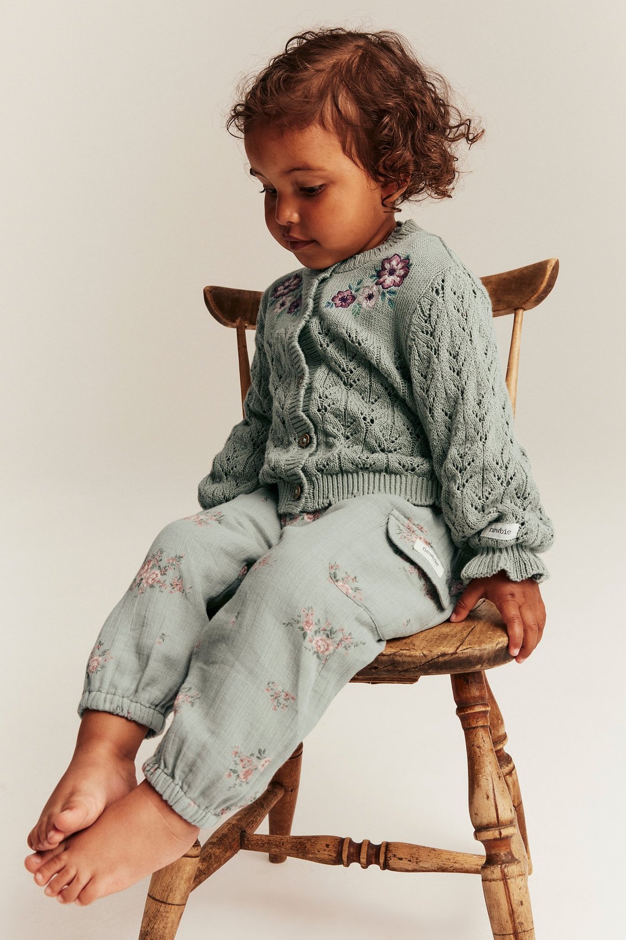 Sidovy av ett barn i gröna blommiga vävda byxor med fickor och en ljusgrön stickad kofta.