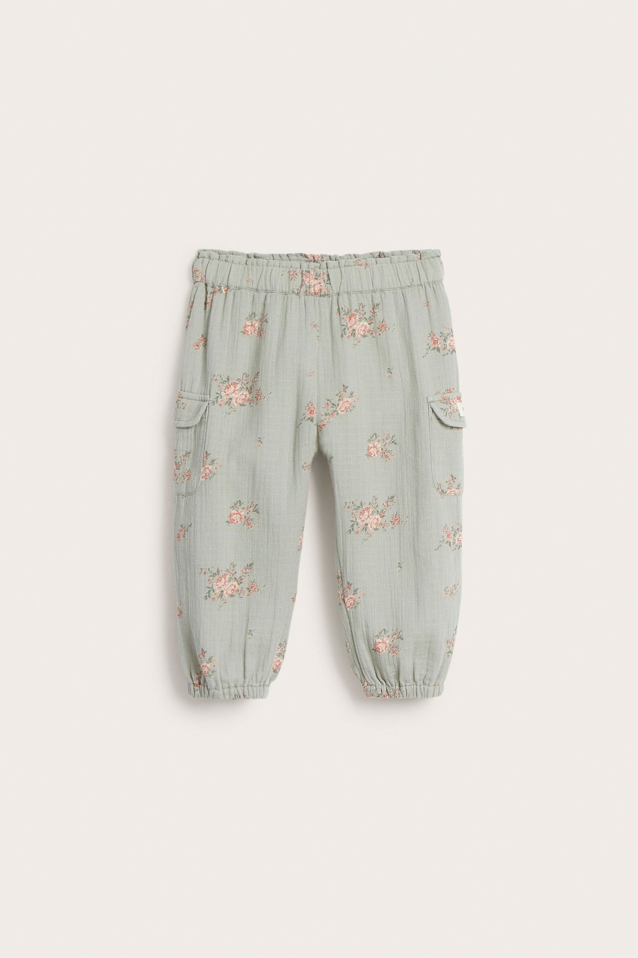 Widok z przodu: Dziecięce jasnozielone spodnie z bawełny w kwiaty, z elastyczną talią, mankietami i kieszeniami z klapkami.
