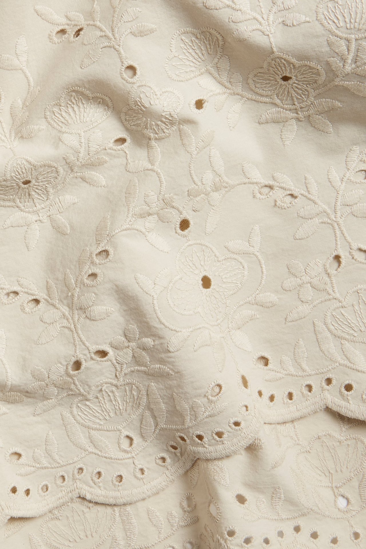 Nærbilde av beige bomullsskjørtstoff med intrikate blomsterbroderier og kamskjellformet hullbroderikant.