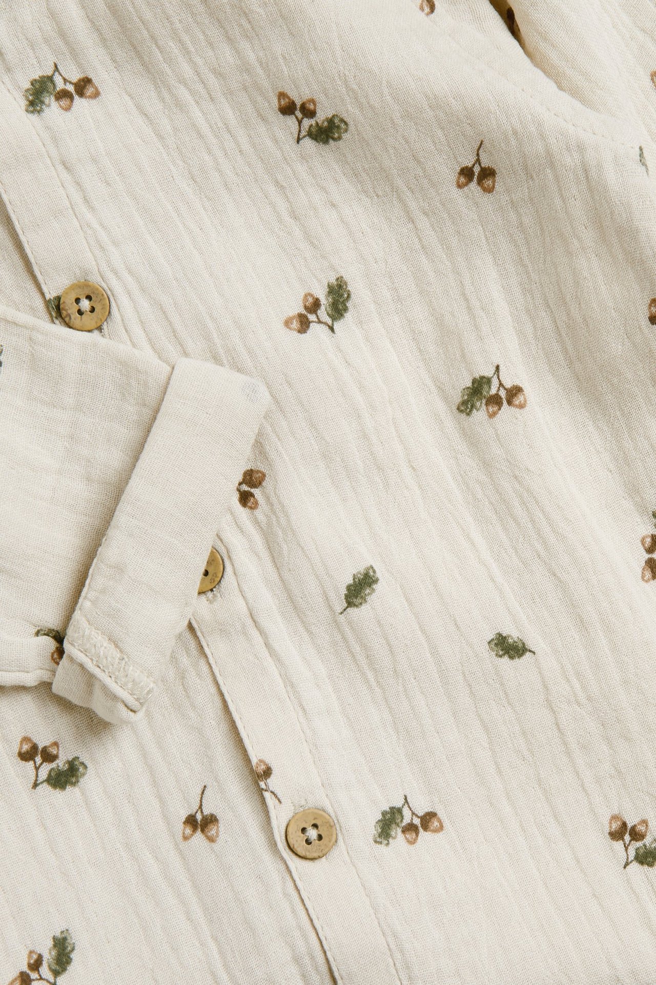 Zbliżenie beżowego kombinezonu z bawełnianego muślinu we wzór żołędzi, z drewnianymi guzikami i podwiniętym mankietem.