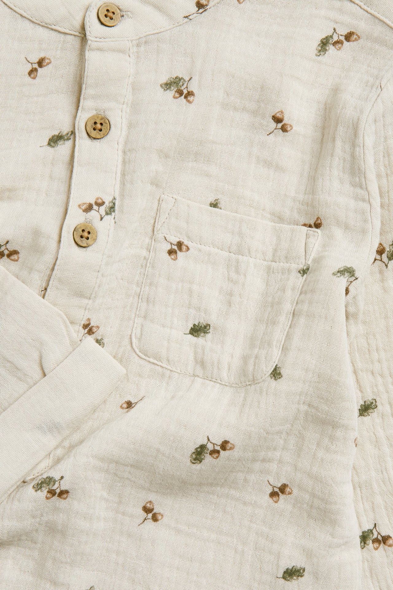 Närbild framifrån: beige långärmad skjorta i bomullsmuslin med ekollonmönster, kokosknappar och bröstficka.