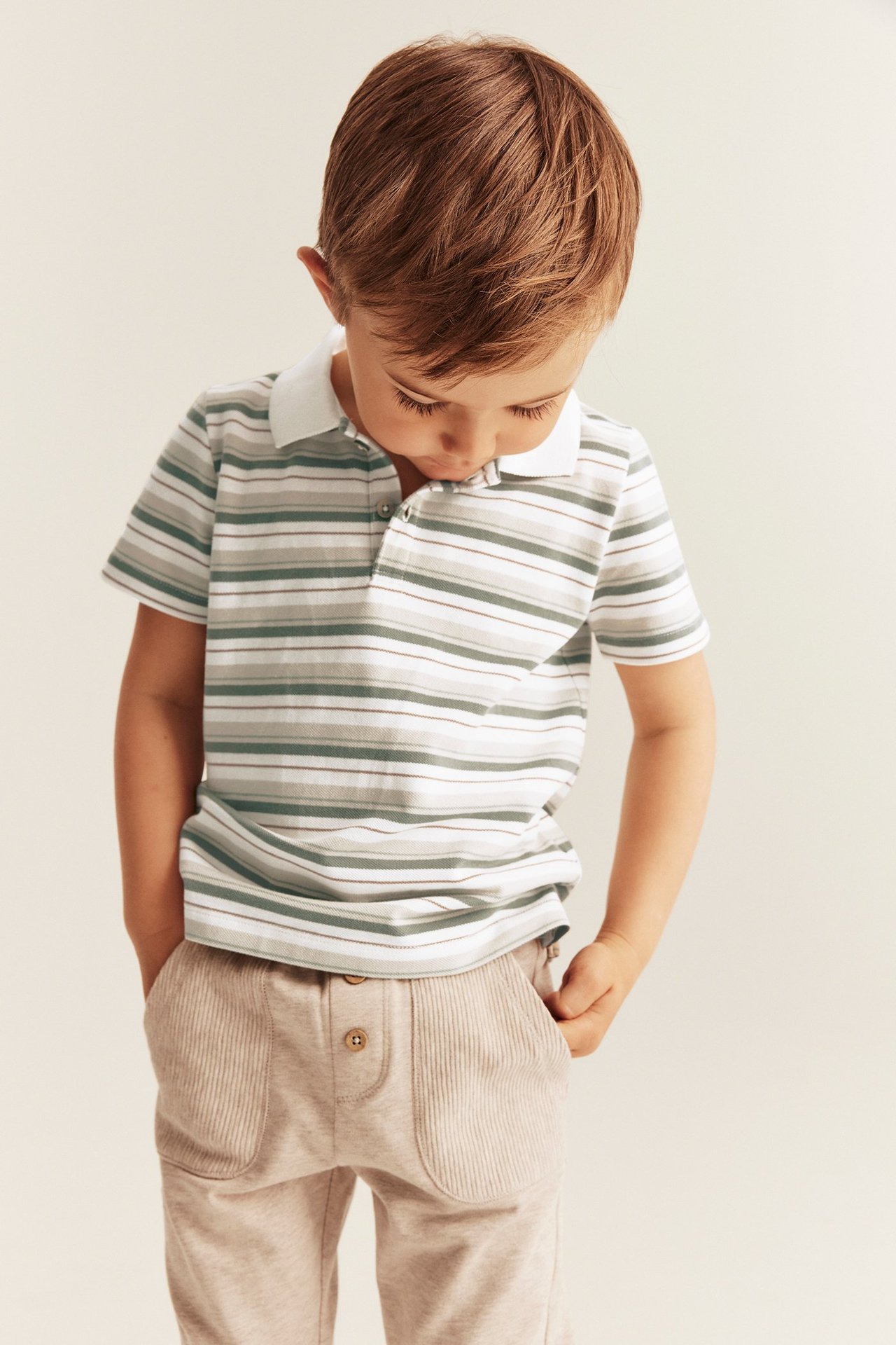 Framifrån, ett barn bär en offwhite, grön- och brunrandig pikétröja i bomull och ljusbeige byxor.