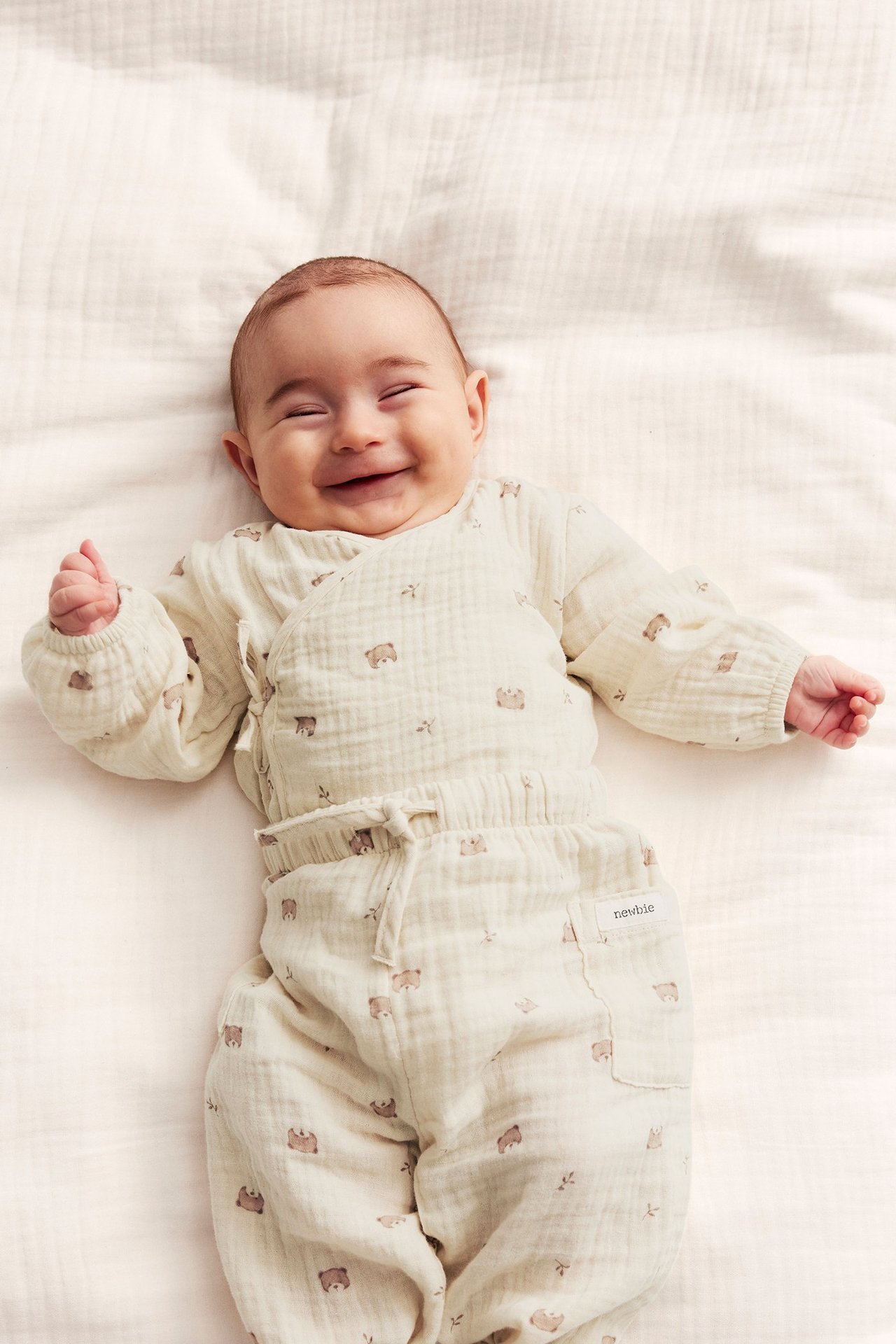 Uppifrån, leende baby i ljusbeige omlottbody och matchande byxor i muslin med nallarmönster.