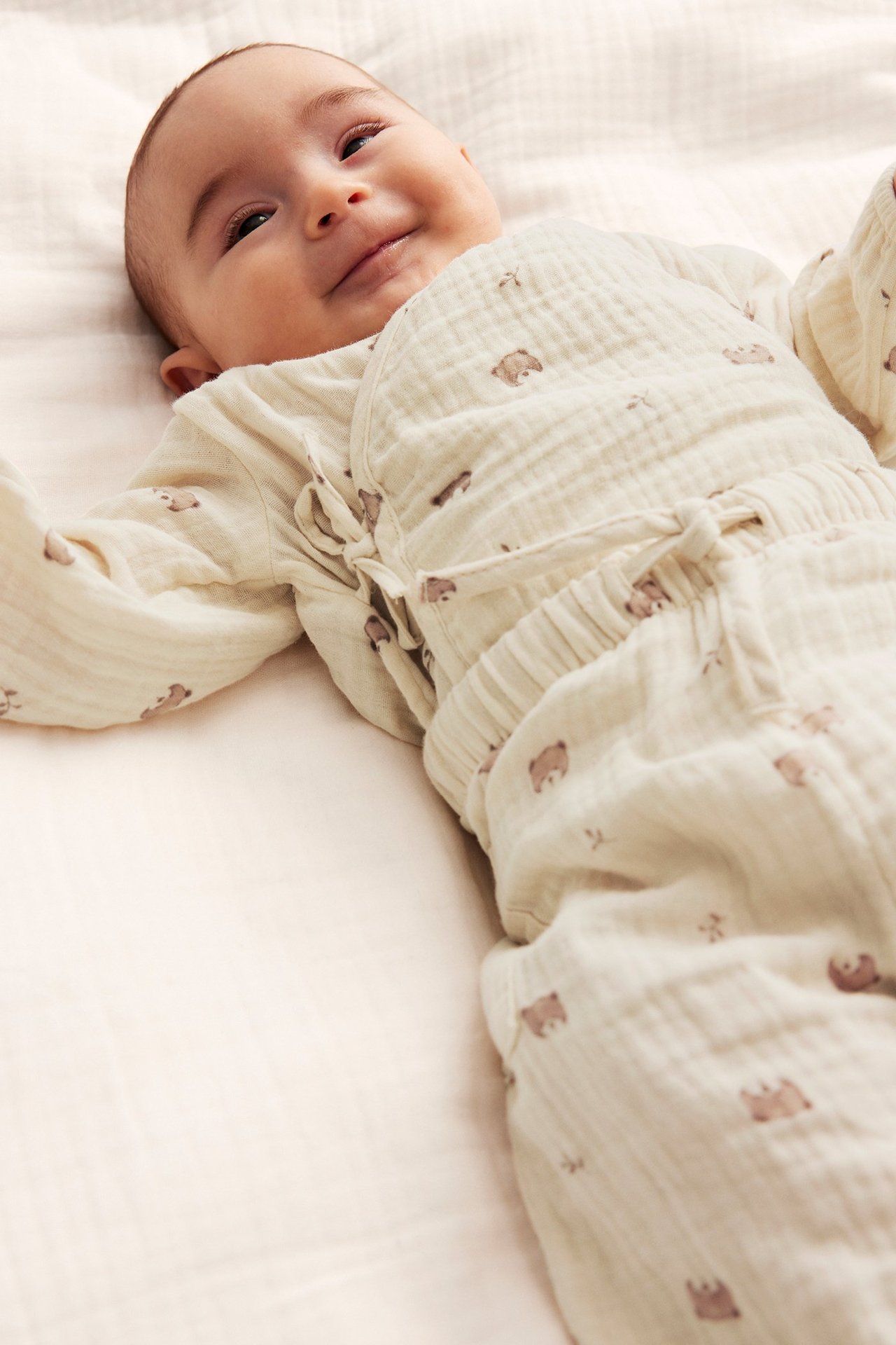 Framifrån: Leende baby i ljusbeige omlottbody och matchande byxor i bomullsmuslin med nallarmönster.