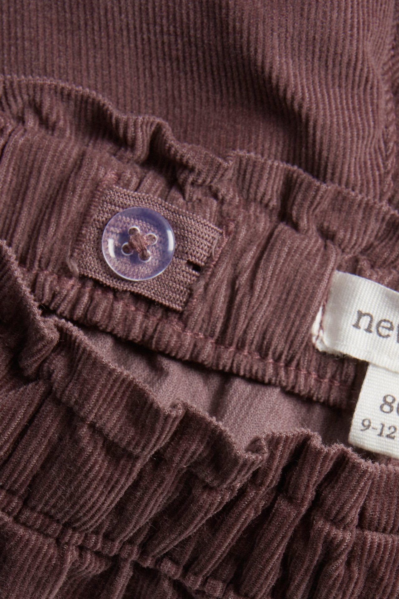 Lähikuva liiloista vakosamettihousuista, joissa säädettävä kuminauhavyötärö ja läpinäkyvä nappi.