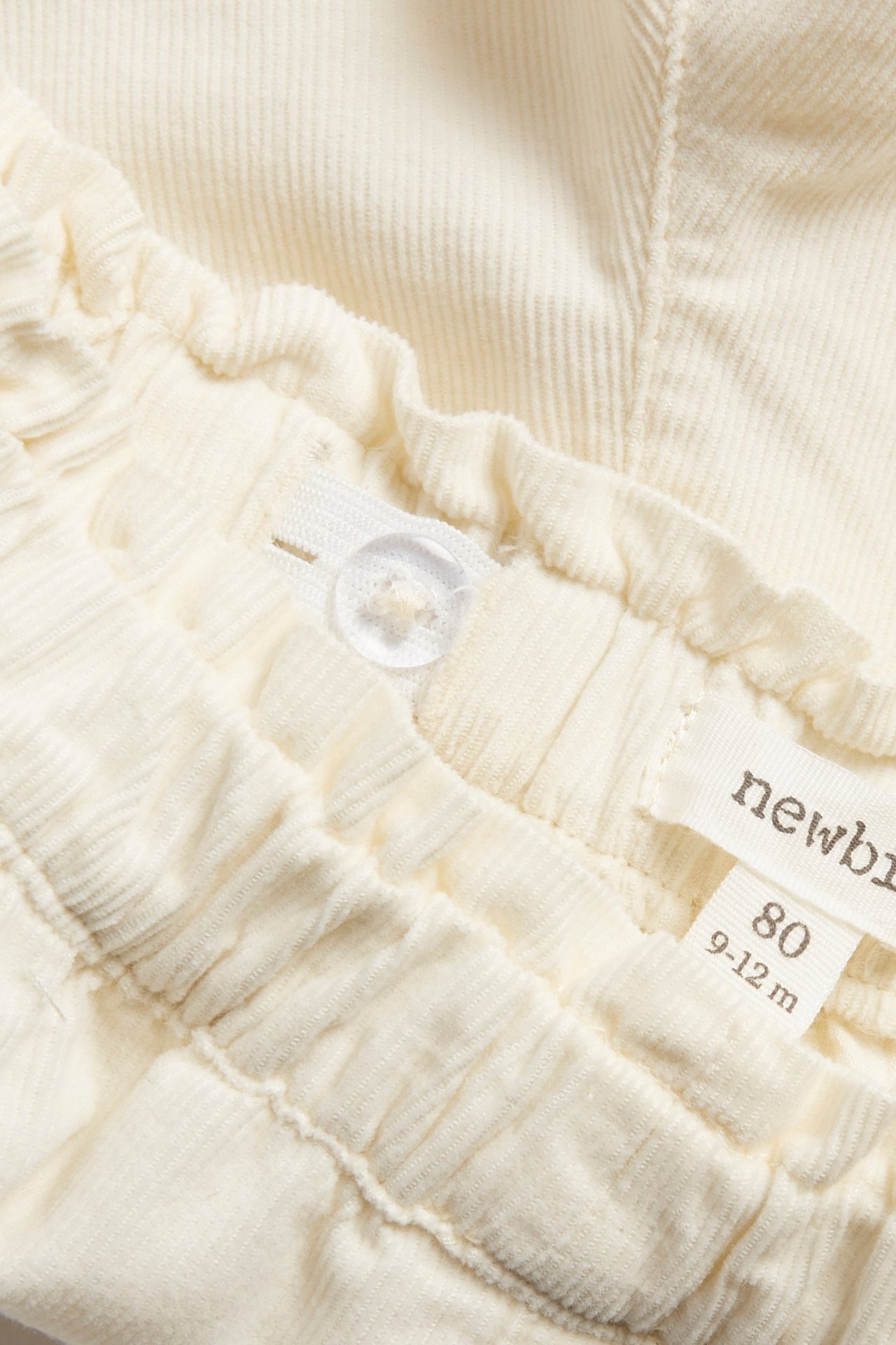 Nærbilde av lysebeige kordfløyelsbukse med elastisk midje og justerbar knapp.