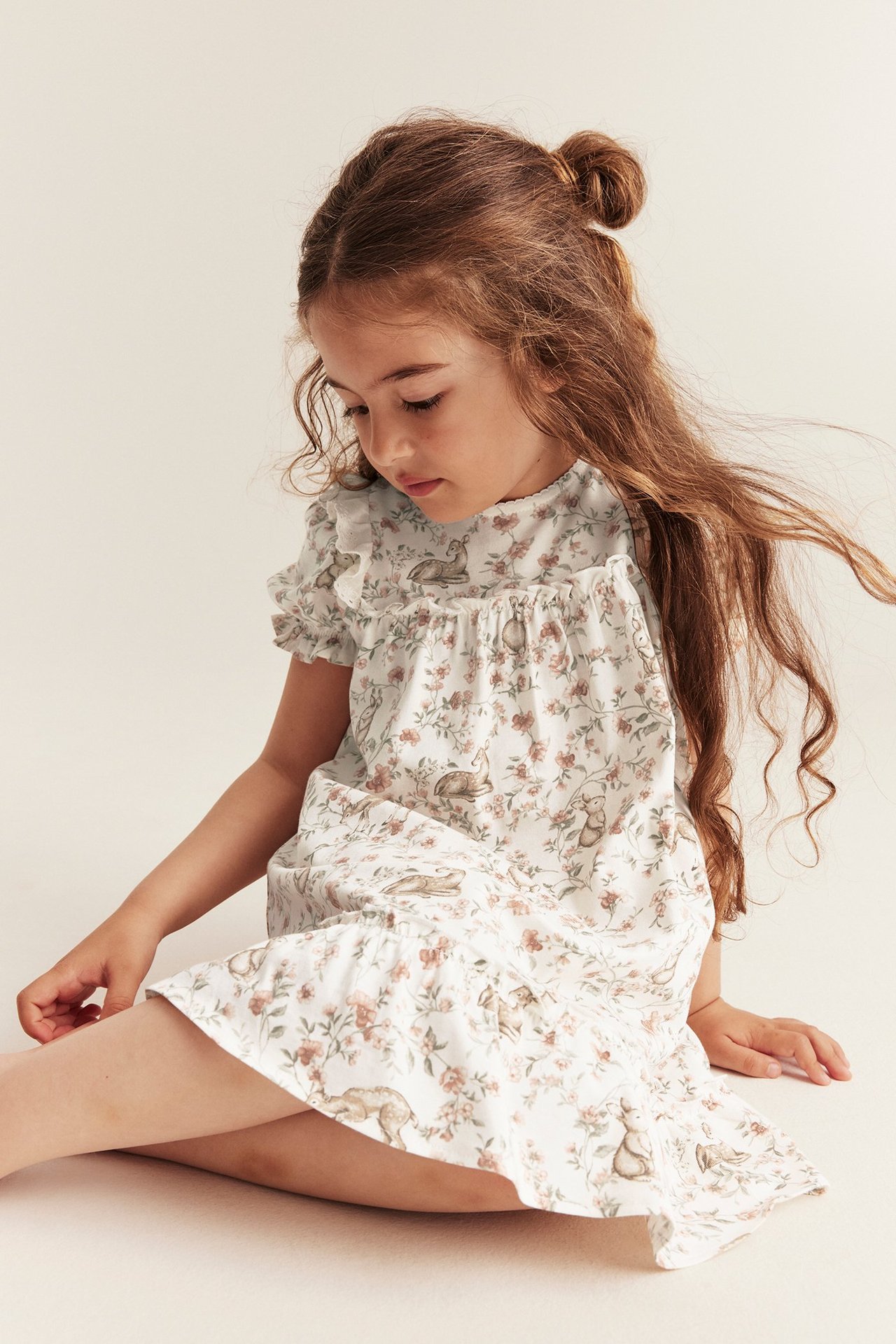Frontvisning av et barn i en offwhite bomullskjole med blomster- og dyremønster, puffermer og volanger.