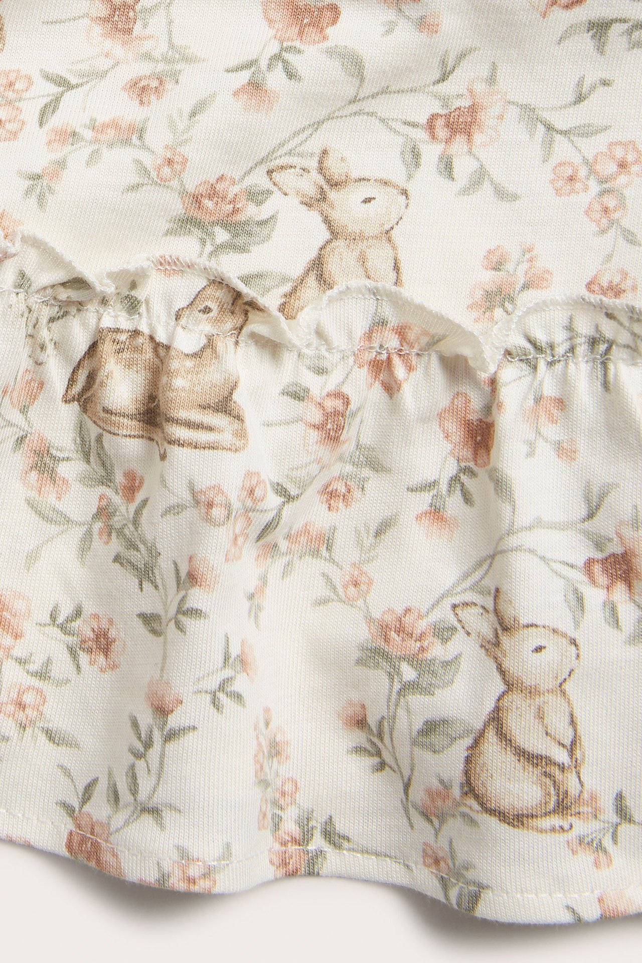 Zbliżenie na ecru bawełnianą tkaninę sukienki z wzorem królików i kwiatów oraz falbanką.