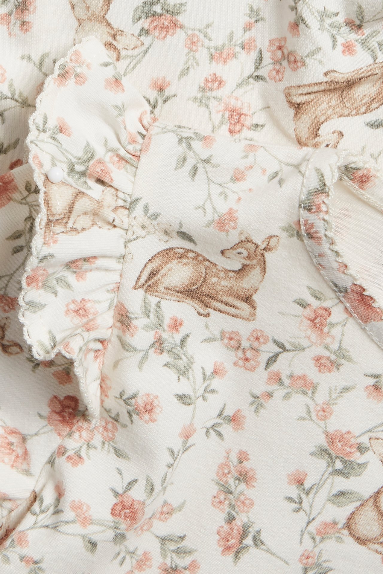 Närbild av en offwhite babybody med blommigt och djurmönster samt volang med picotkant.