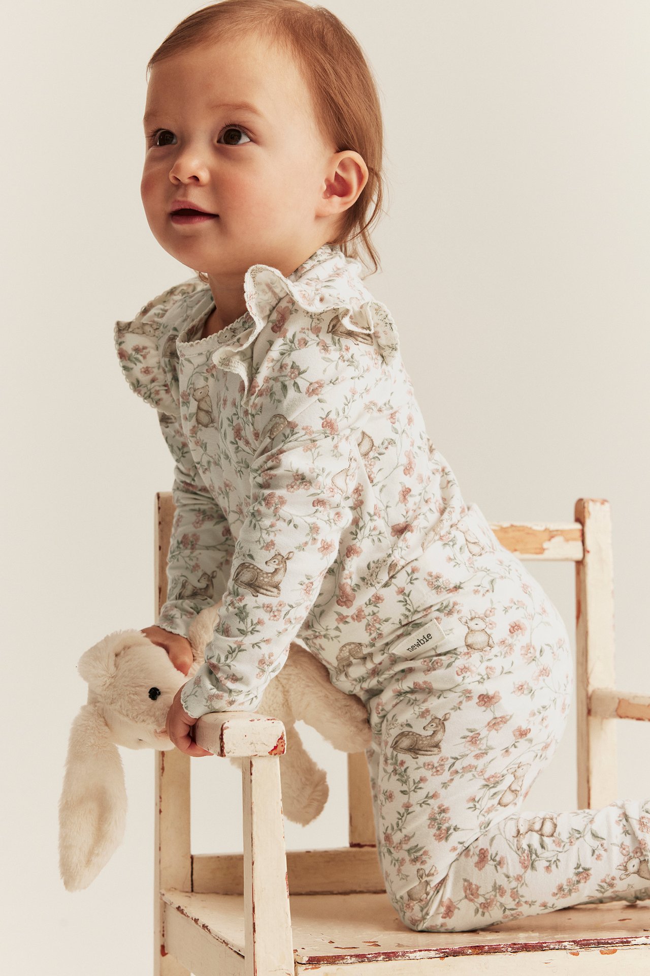 Sned framifrån-vy av baby i offwhite blommönstrad body med djur och volanger, håller vit kanin.