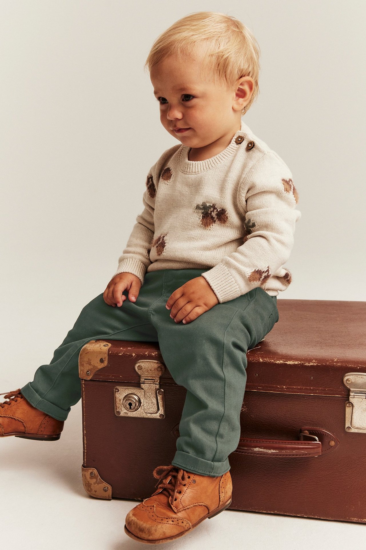 Edestäpäin vauva tummanvihreissä kudotuissa housuissa, beigessä kuvioidussa neuleessa ja ruskeissa nauhakengissä.