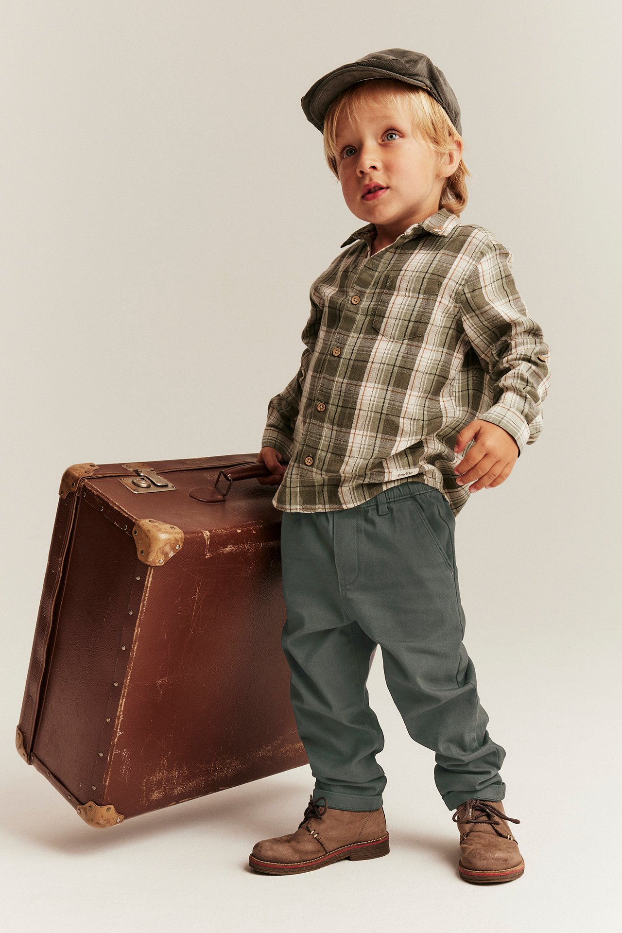 Framifrån, ett barn i mörkgröna vävda byxor, en rutig skjorta, en mörk keps och bruna skor.