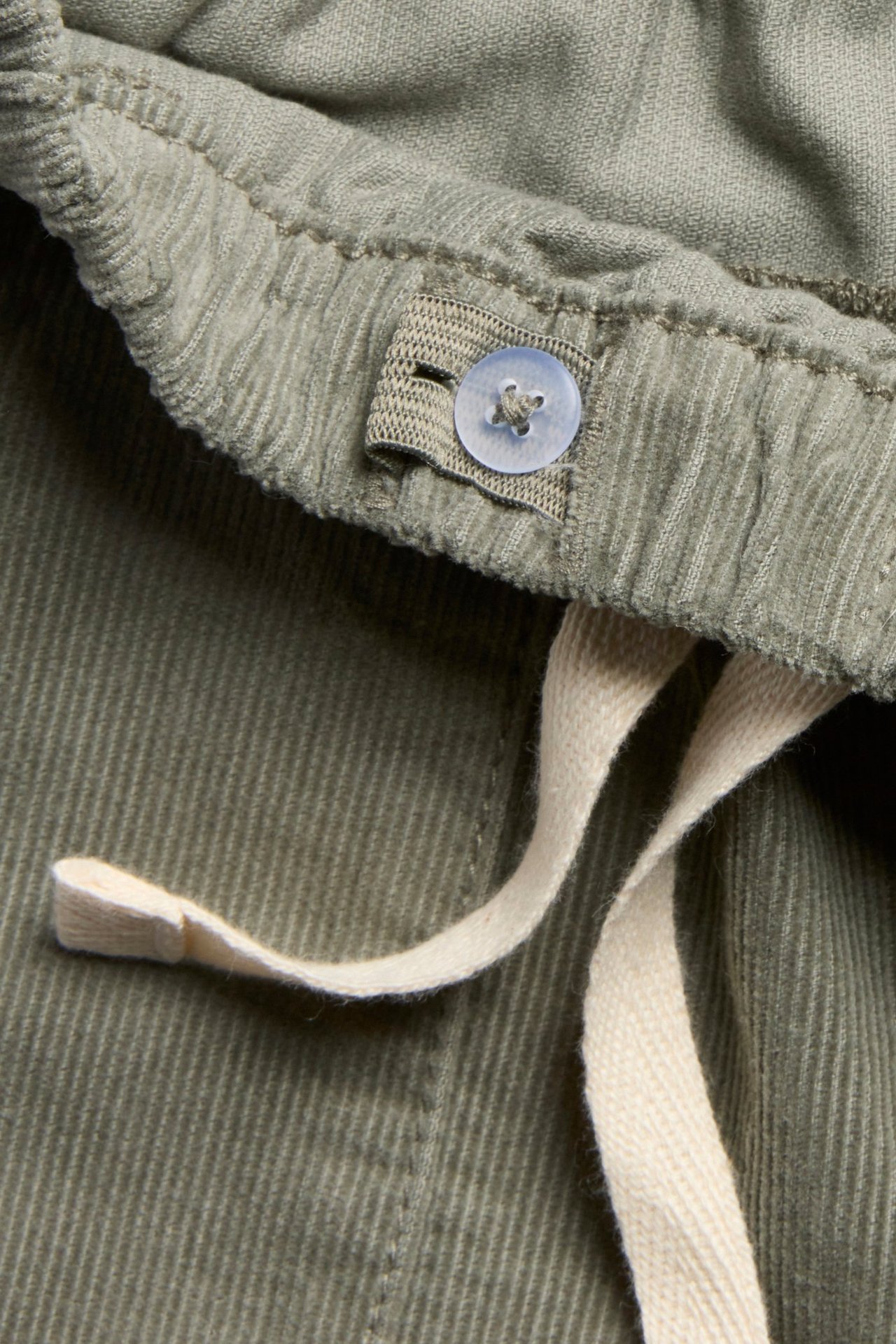 Lähikuva tummanvihreistä vakosamettihousuista: joustava vyötärö, vaaleansininen nappi, kermanvärinen kiristysnyöri.