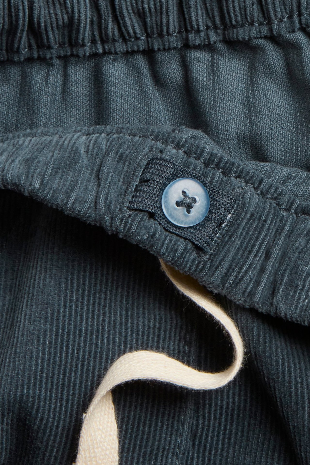 Lähikuva tummansinisistä vakosamettihousuista, joissa joustava vyötärö, vaaleansininen nappi ja beige nyöri.