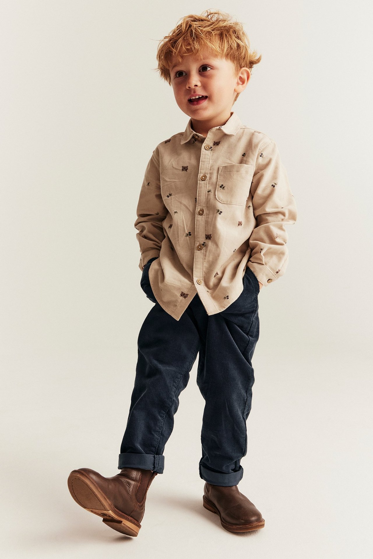 Edestäpäin, lapsi tummansinisissä vakosamettihousuissa, beigessä kuviollisessa paidassa ja ruskeissa saappaissa.