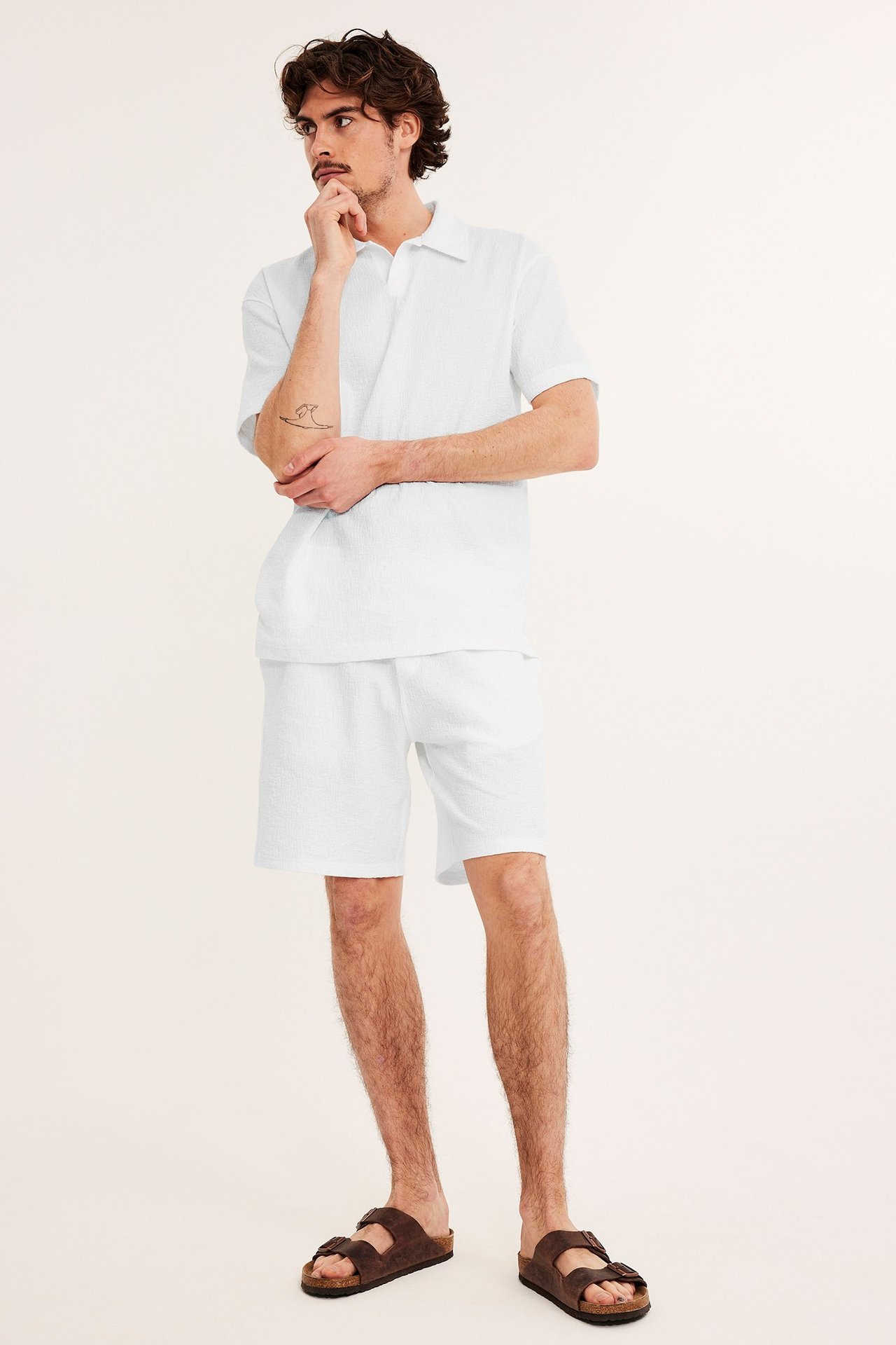 Edestä: mies yllään luonnonvalkoiset kuvioidut shortsit, yhteensopiva pikeepaita ja ruskeat sandaalit.